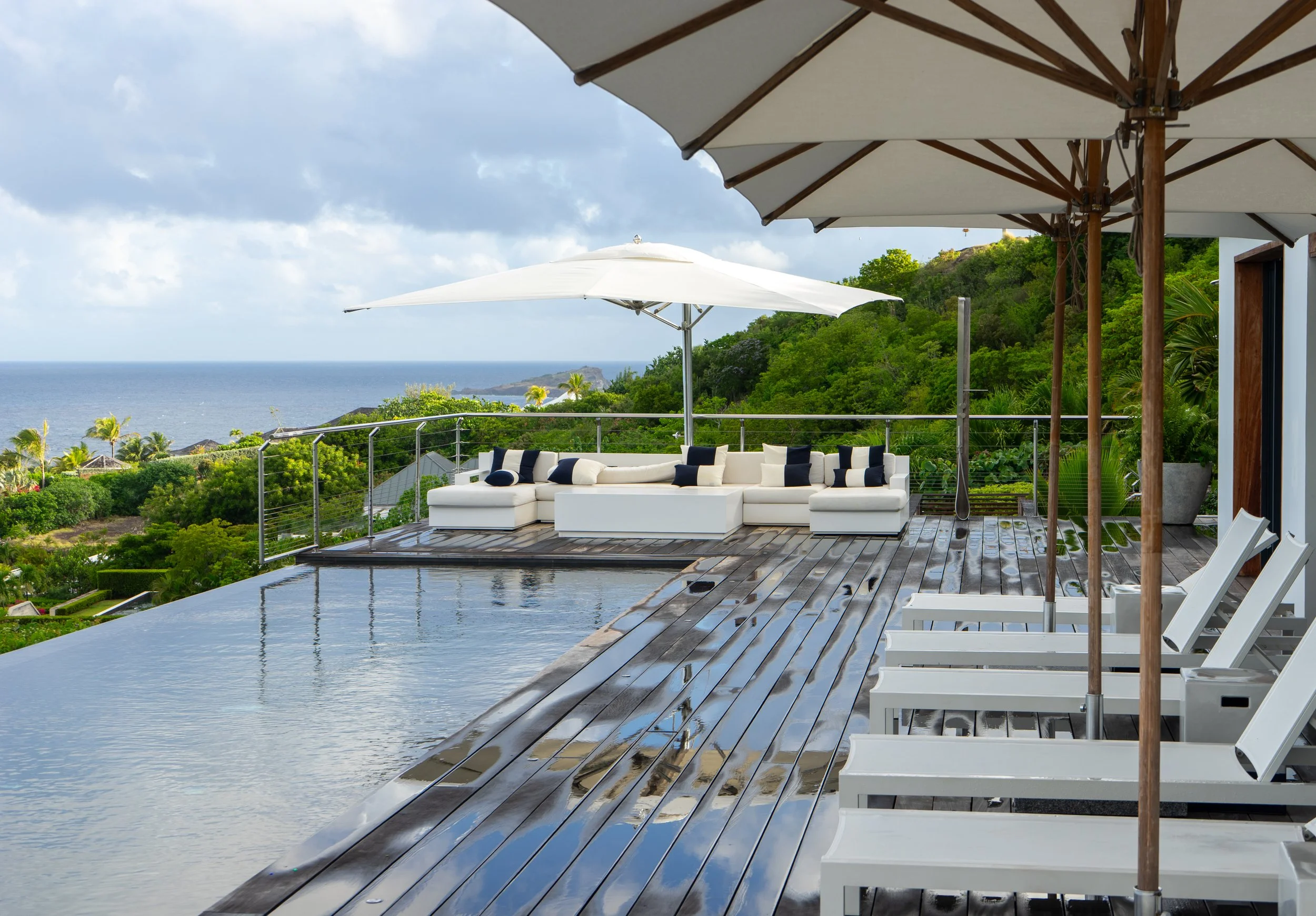 A luxurious outdoor patio with a swimming pool, white seating area with pillows, large white umbrellas providing shade, surrounded by lush green hills and a view of the ocean in the distance.