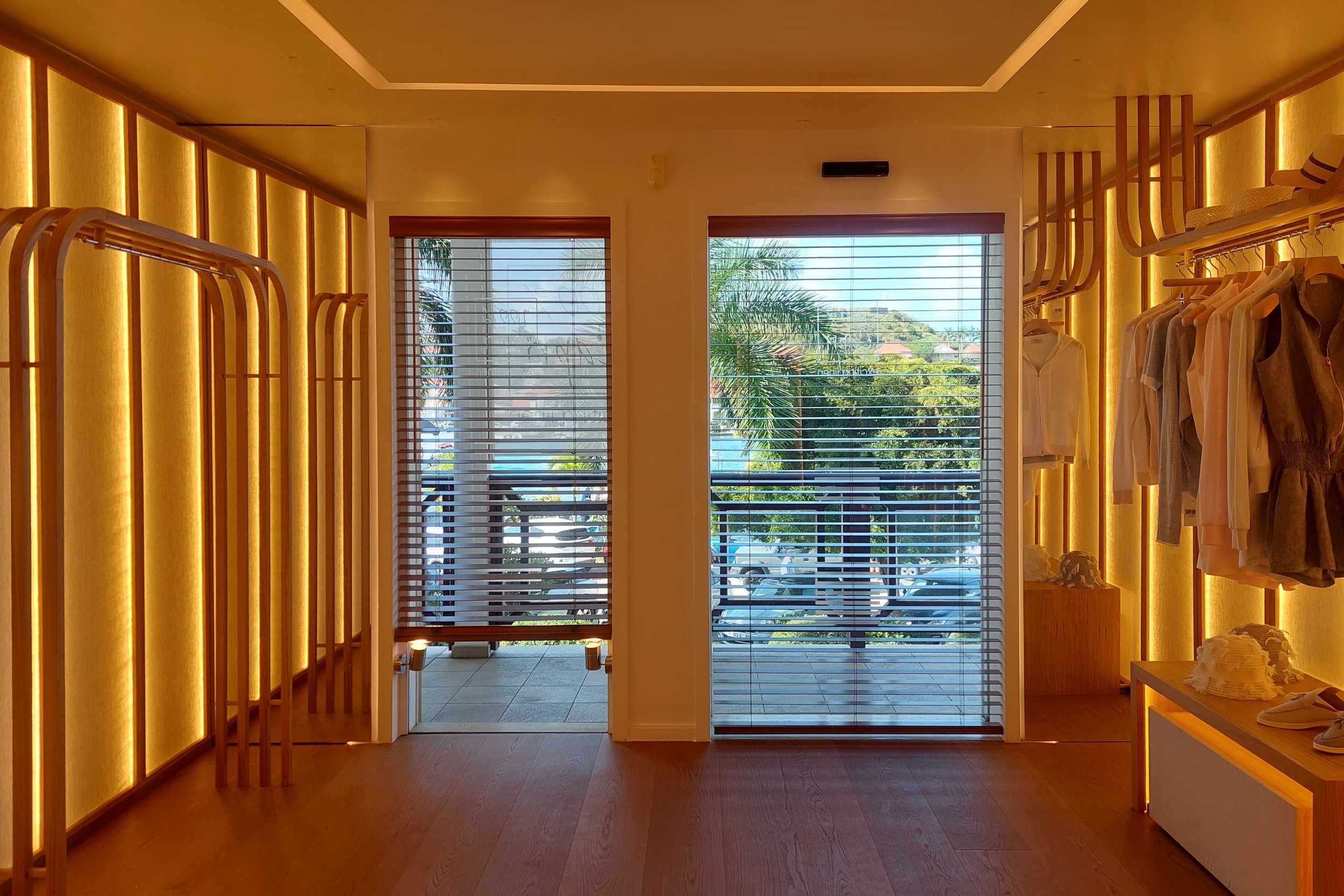 Magasin de vêtements intérieur avec des présentoirs en bois, une grande fenêtre offrant une vue sur des palmiers et un parking à l’extérieur, ainsi qu’une porte donnant accès à l’extérieur.