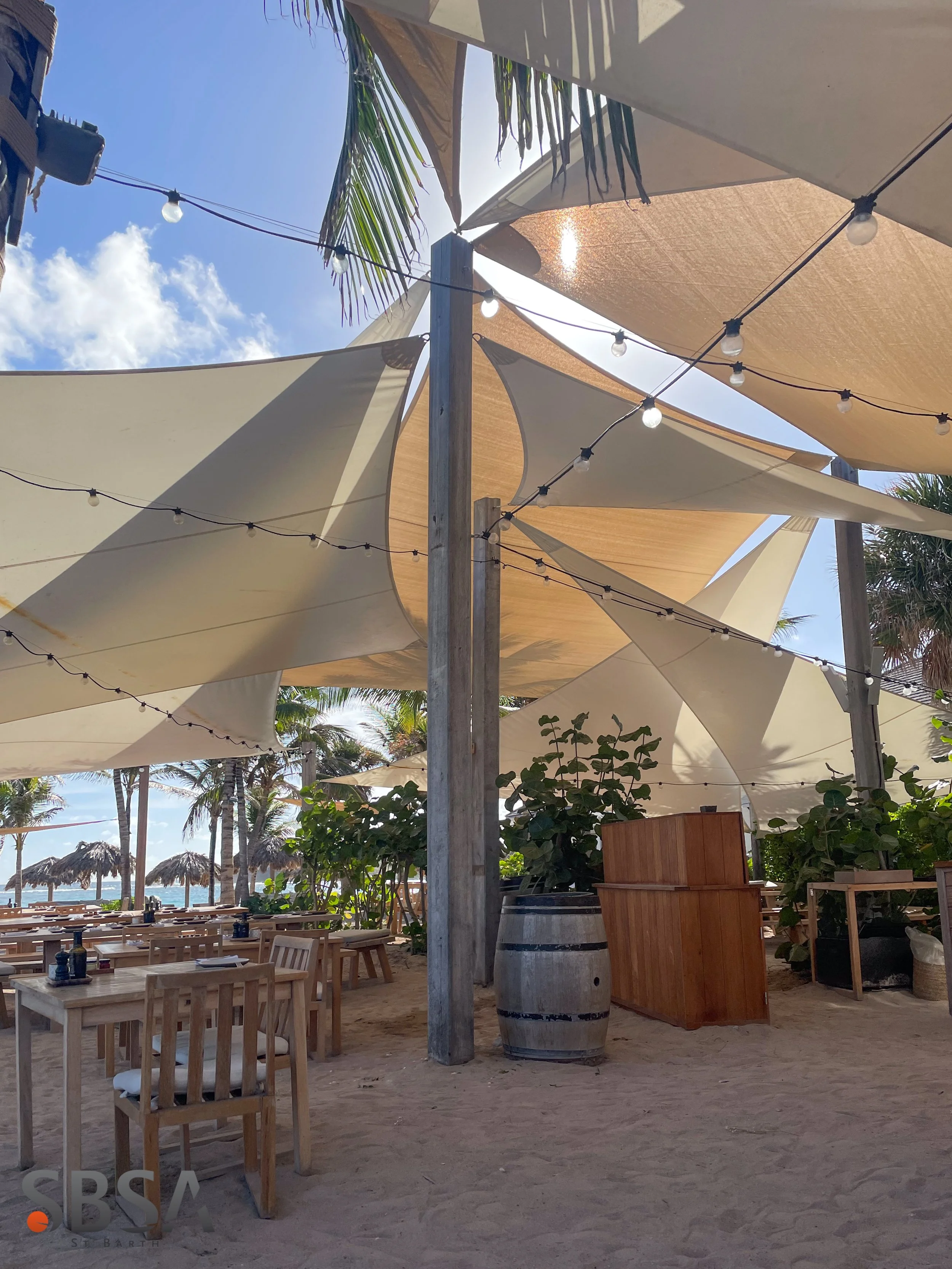 Outdoor beach restaurant with wooden tables and chairs, large tent-like shades, string lights, and palm trees in the background under a bright blue sky.