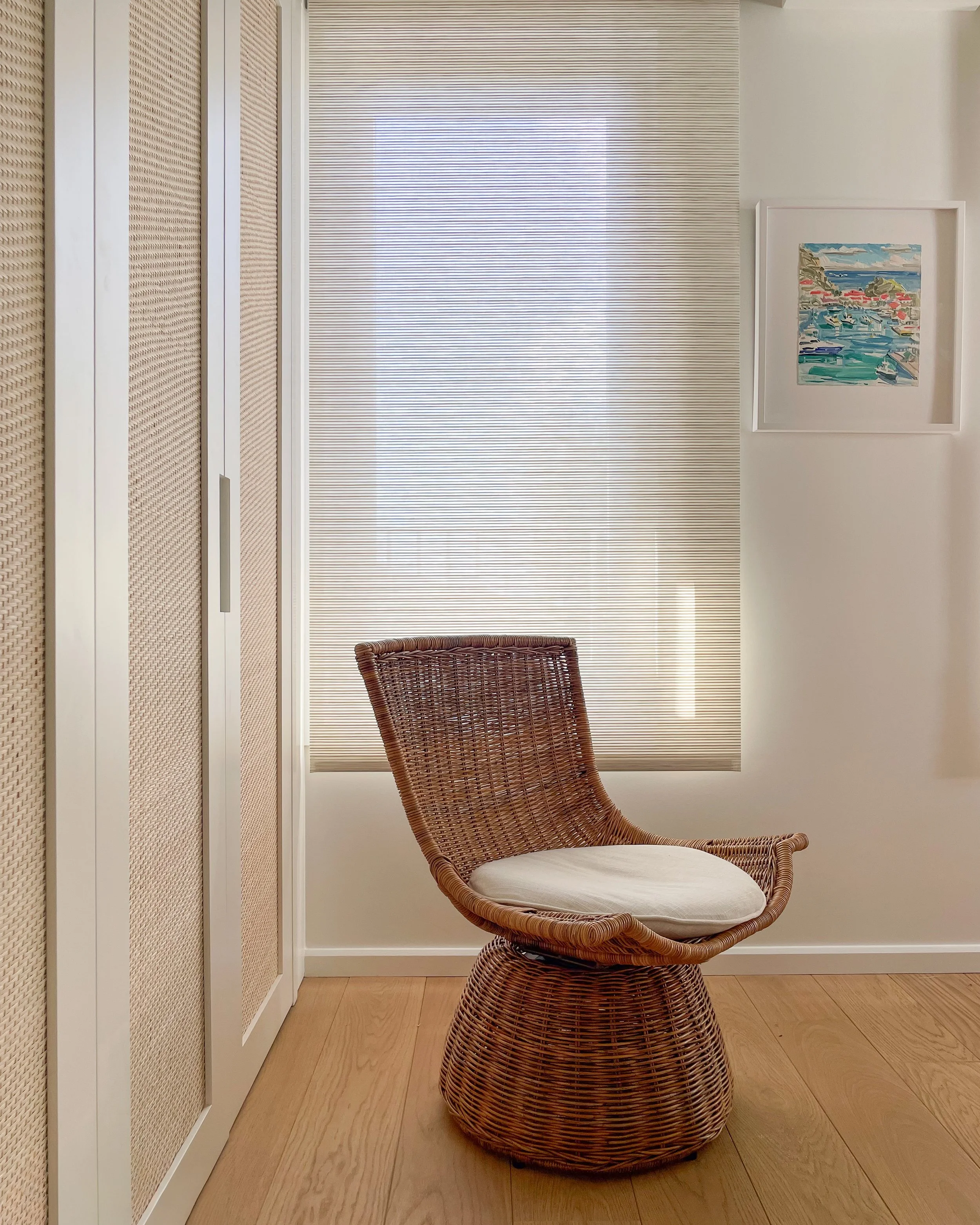 Une chaise en osier avec un coussin blanc dans une pièce lumineuse au style minimaliste. Derrière la chaise se trouve une grande fenêtre équipée de stores horizontaux, ainsi qu’un tableau encadré aux couleurs vives représentant une scène côtière. À g