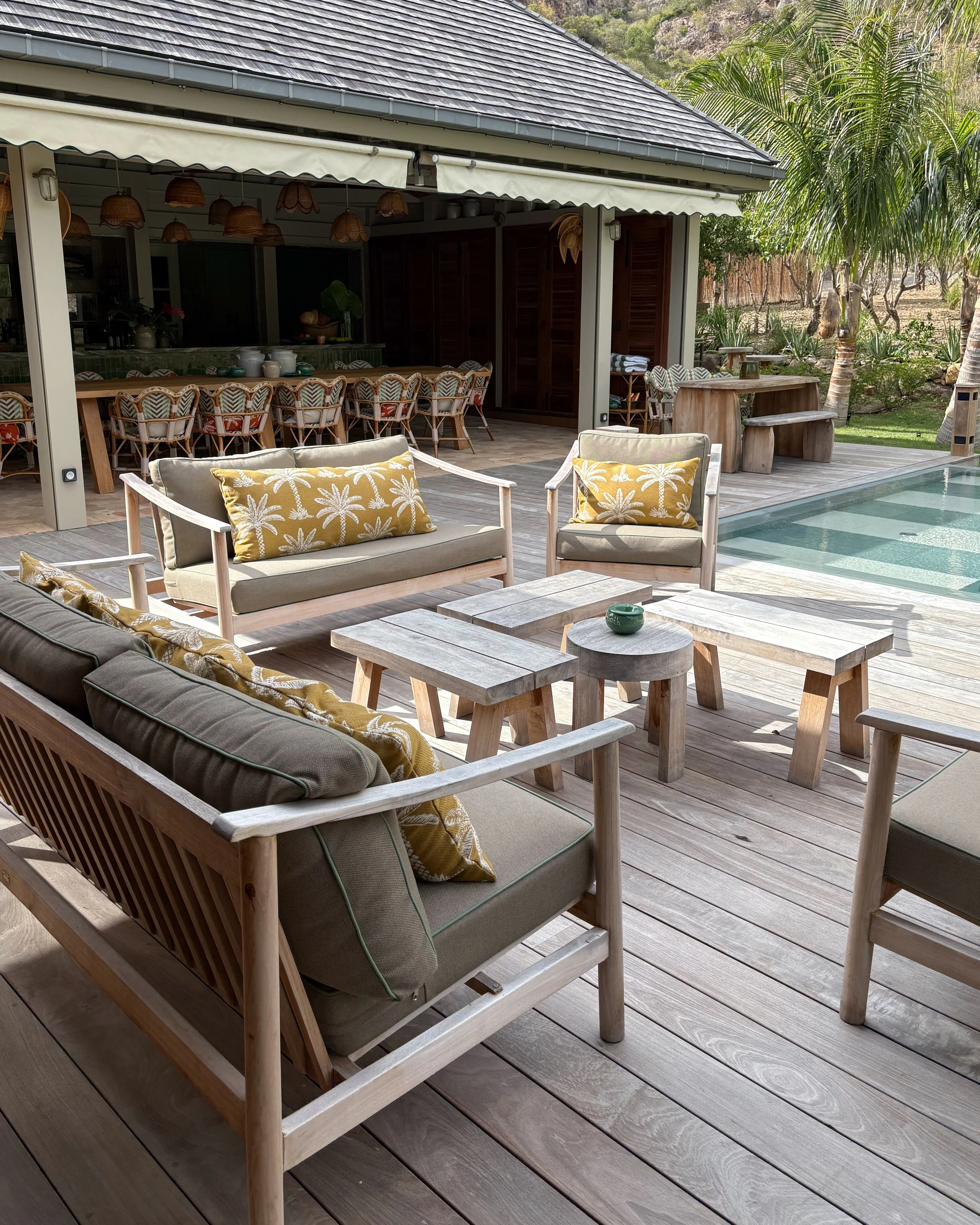 Outdoor patio with wooden furniture, including a beige couch with decorative pillows, a wooden coffee table, and a bench. There is a pool on the right side with water, trees, and green plants in the background.