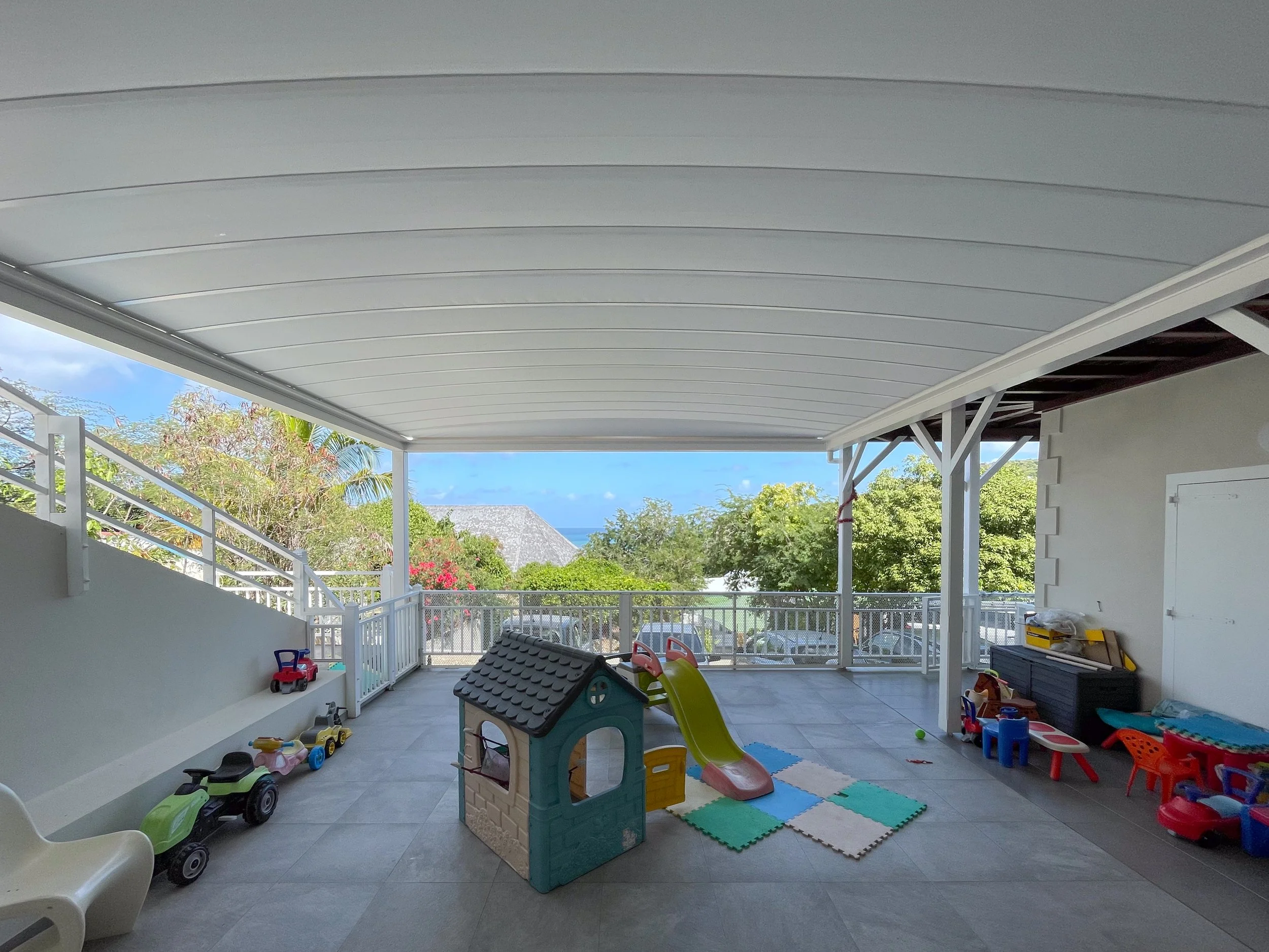 Children's outdoor play area with toys, playhouse, slide, and colorful play mats on a patio with a view of trees and the sky.