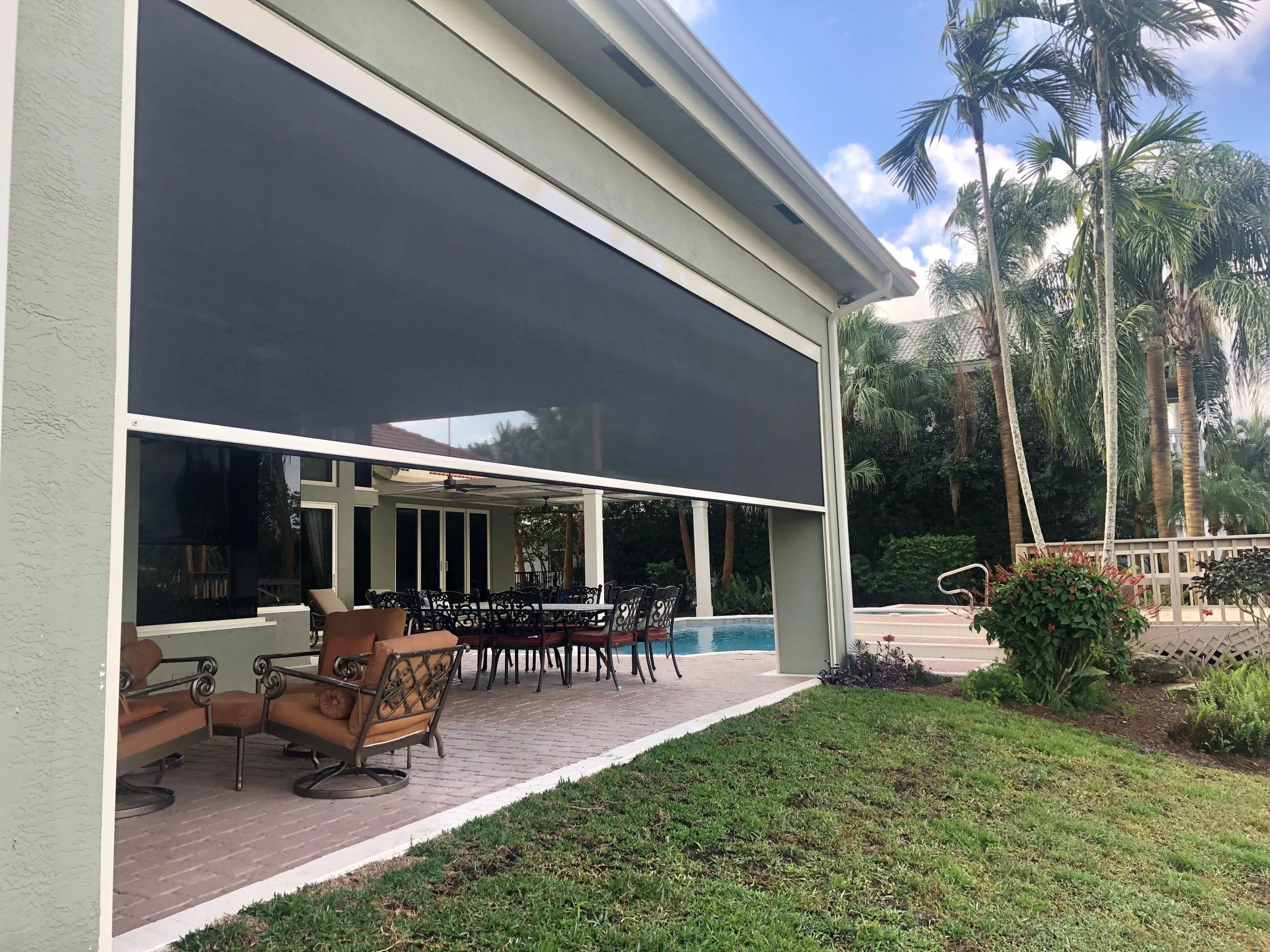 Backyard patio area with outdoor seating, including cushioned chairs and a table, adjacent to a swimming pool. The patio is under a screened porch with a black shade and surrounded by lush palm trees and greenery, with a house and a blue sky with clo