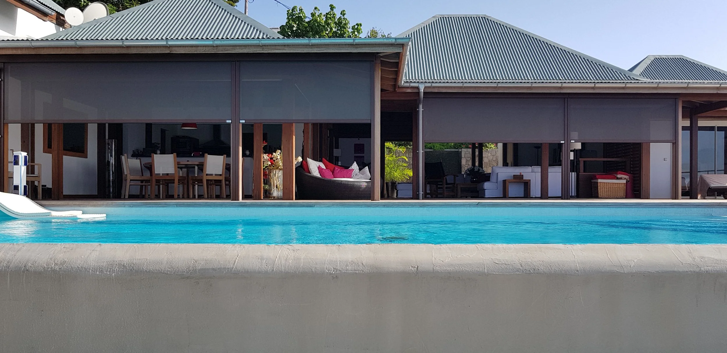View of a backyard area with a swimming pool in the foreground, and a modern house with multiple electric vertical stores, outdoor furniture, and a corrugated metal roof in the background.