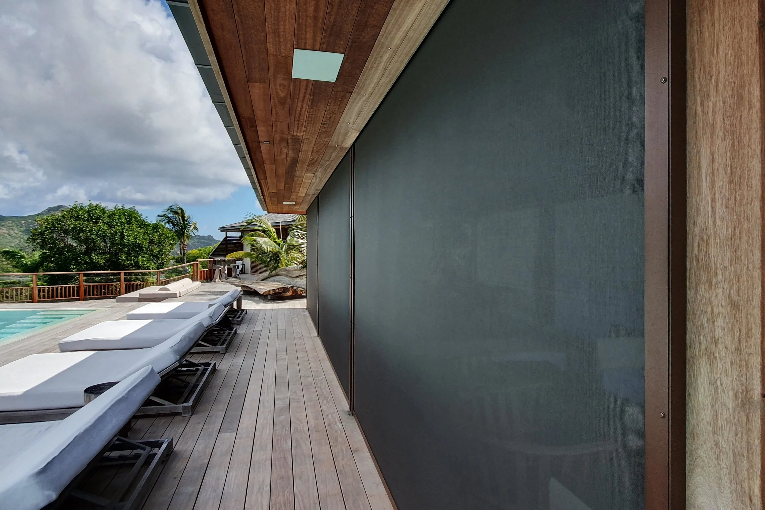 A modern outdoor patio with lounge chairs, a swimming pool, lush green trees, palm trees, mountains in the background, and a wooden building with a sloped roof and a wooden wall with a small window.