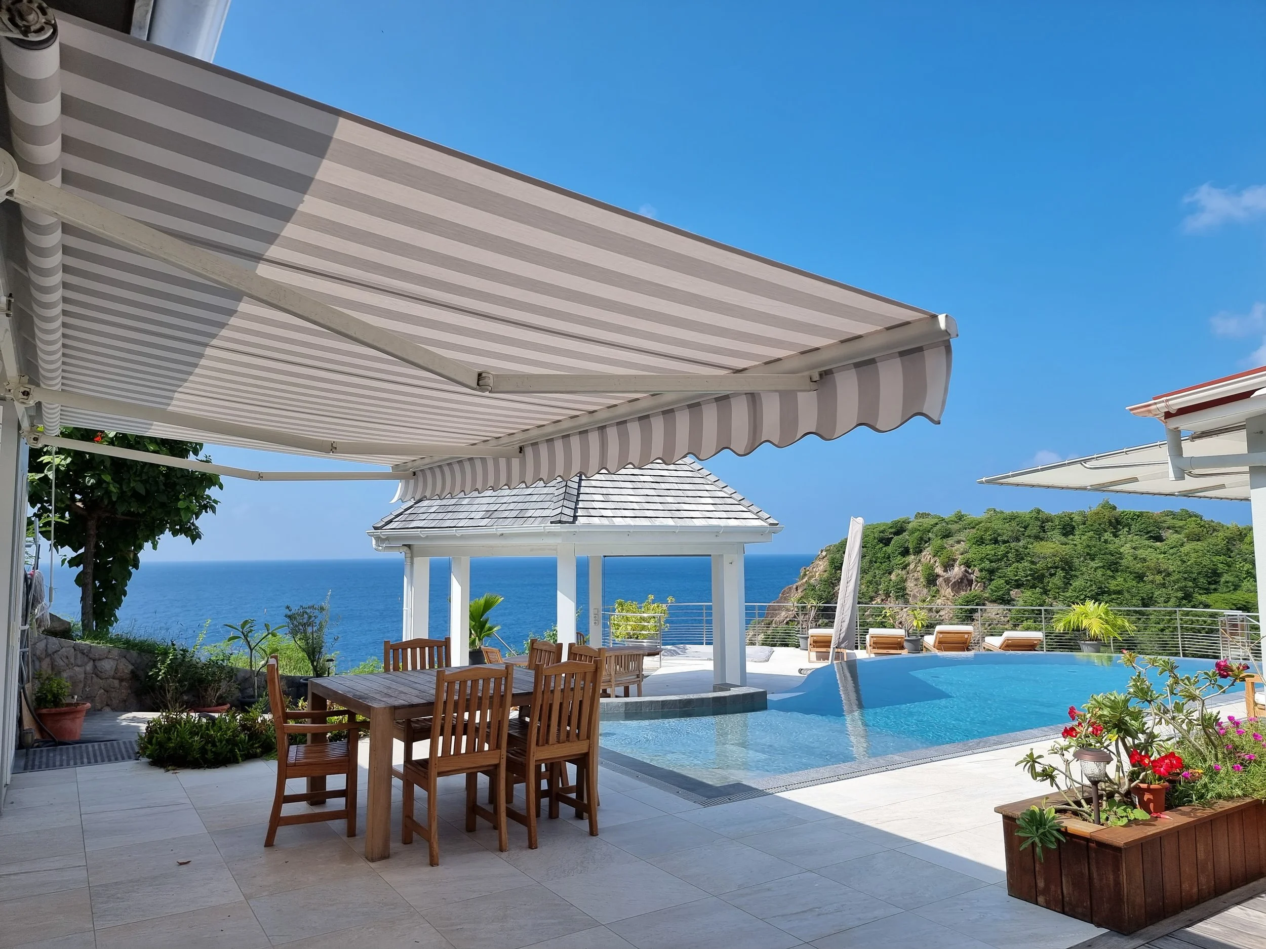 Outdoor patio area with a striped retractable awning, wooden dining table with chairs, a swimming pool, potted plants, and a scenic ocean view in the background.