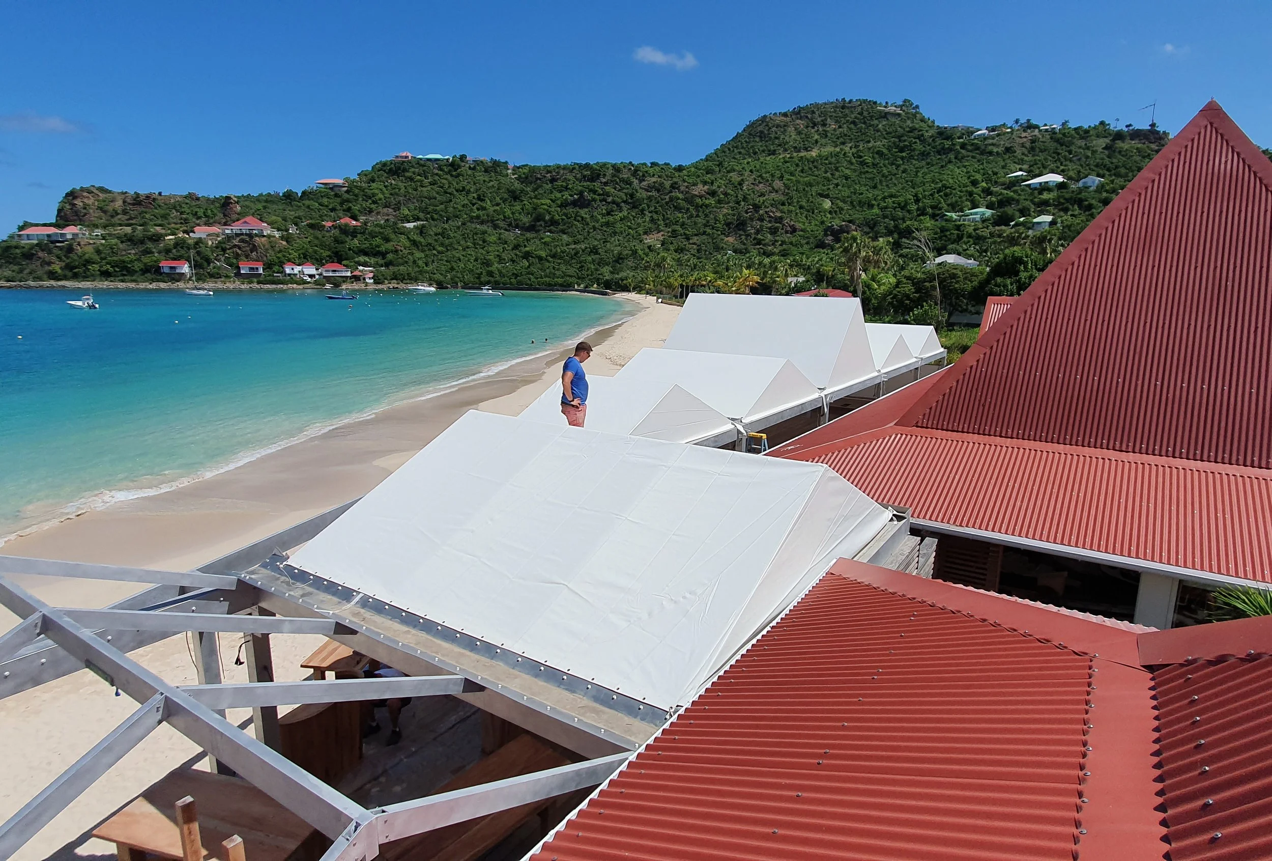 A man stands on a balcony overlooking a white sandy beach, turquoise water, and lush green hills with small houses and structures on a sunny day.