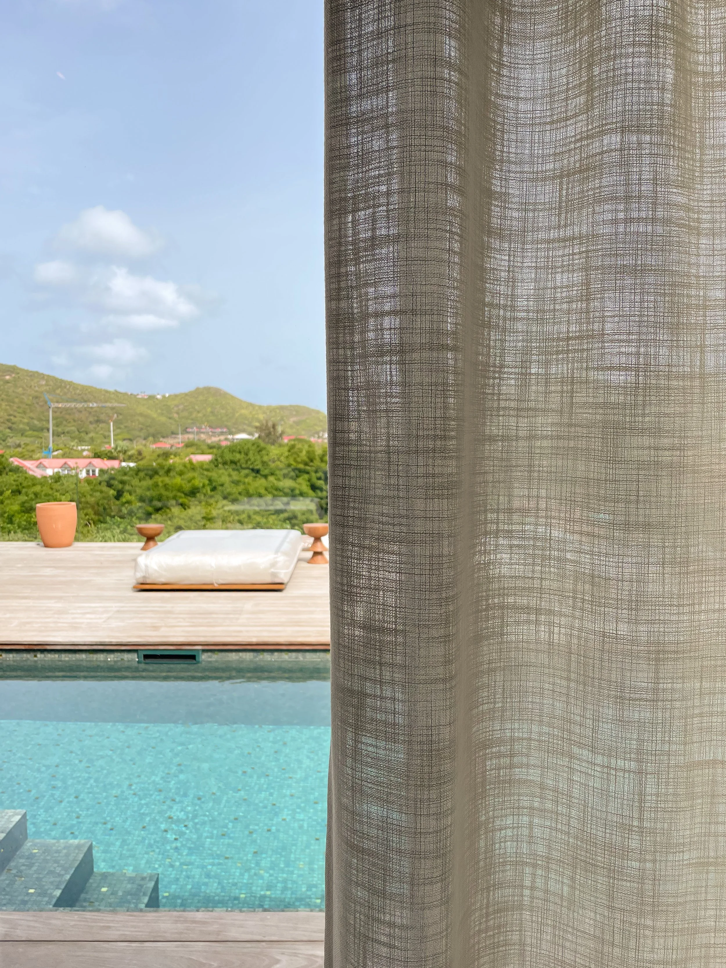 Vue d'une piscine extérieure avec de l'eau turquoise, entourée d'une terrasse en bois, avec un volet en tissu léger entre la piscine et la vue sur un paysage vallonné avec des maisons et des arbres, sous un ciel bleu avec quelques nuages.
