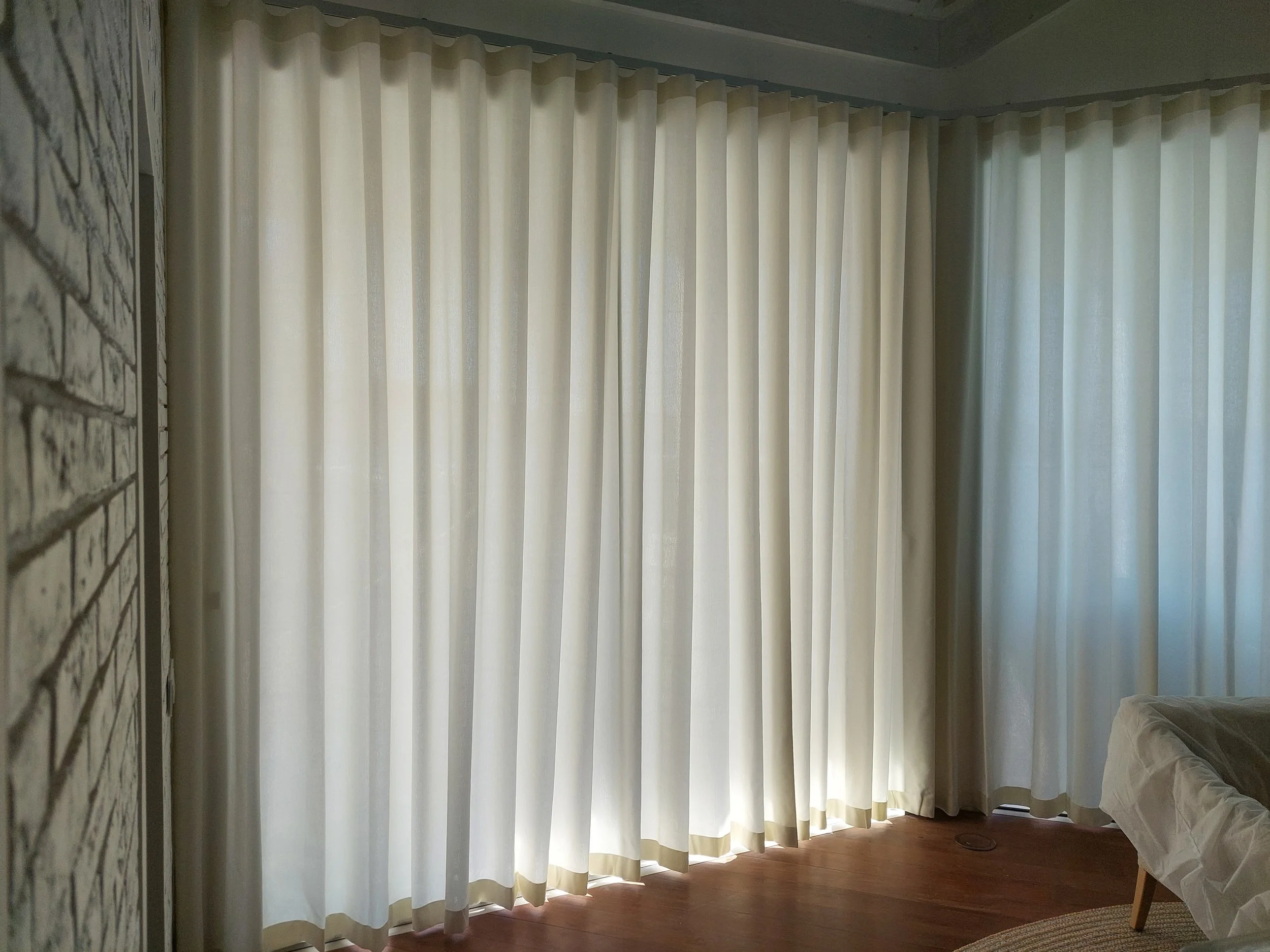 Cream-colored curtains hanging over a large window in a room. The room has white walls, a wooden floor, and a partially visible bed with white bedding and a white armchair.