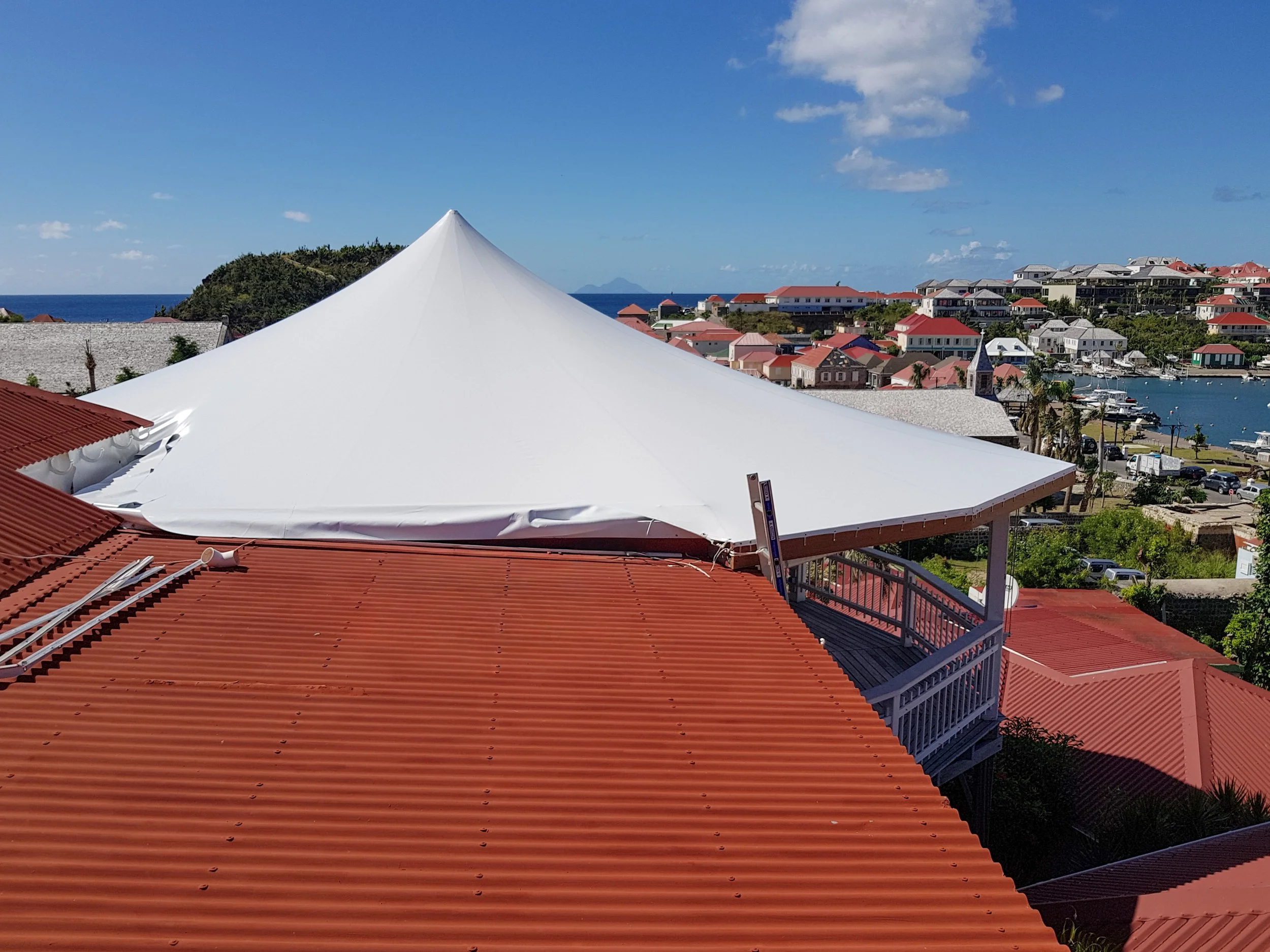 A view of a coastal town with colorful buildings, a marina with boats, and the ocean in the background under a partly cloudy sky. The foreground features a large, white, pointed tent and red-tiled rooftops.