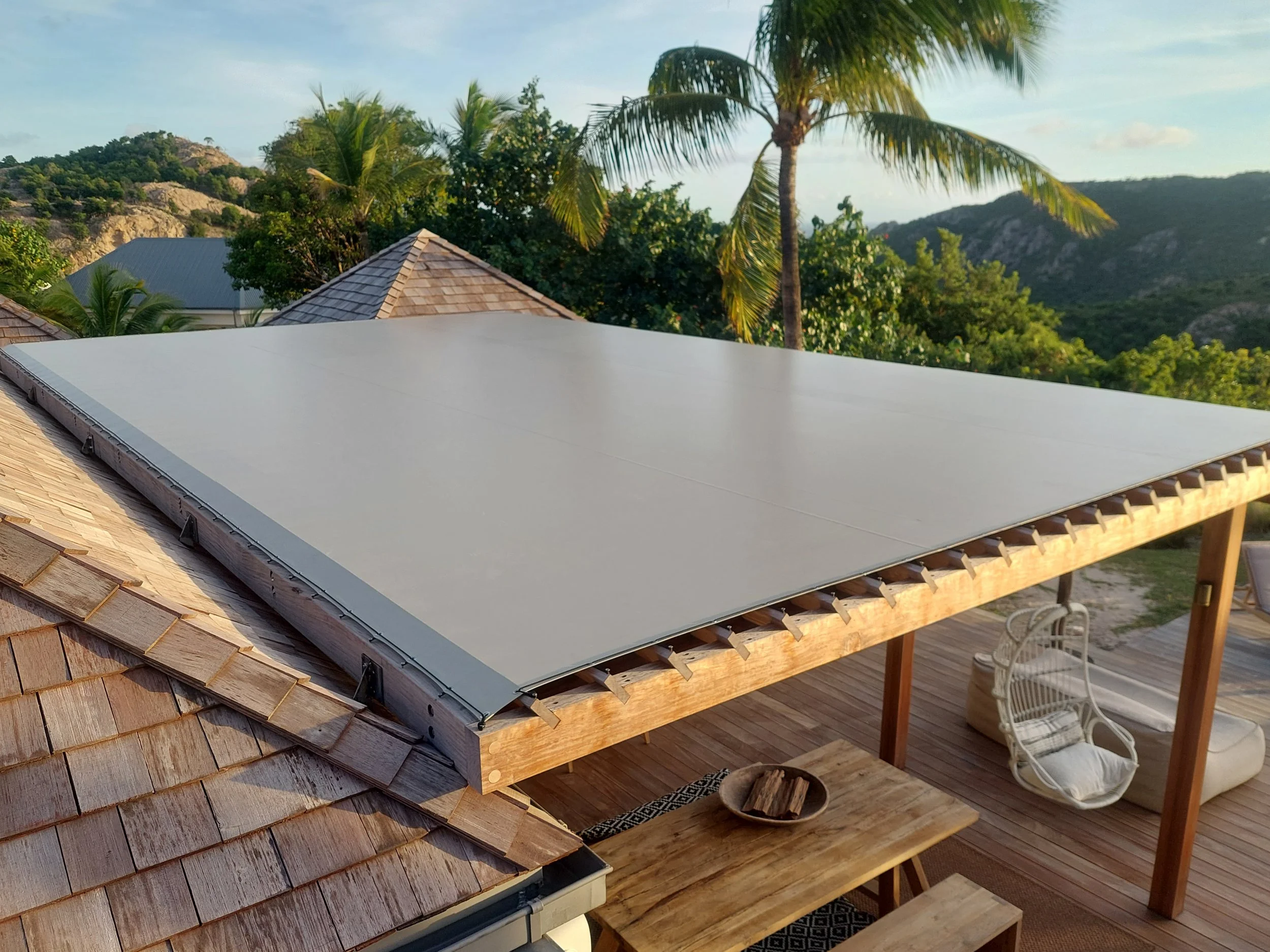 View from a rooftop deck with a solar panel in shadow, surrounded by wooden shingles, wooden flooring, chairs, trees, and distant hills under a blue sky with clouds.