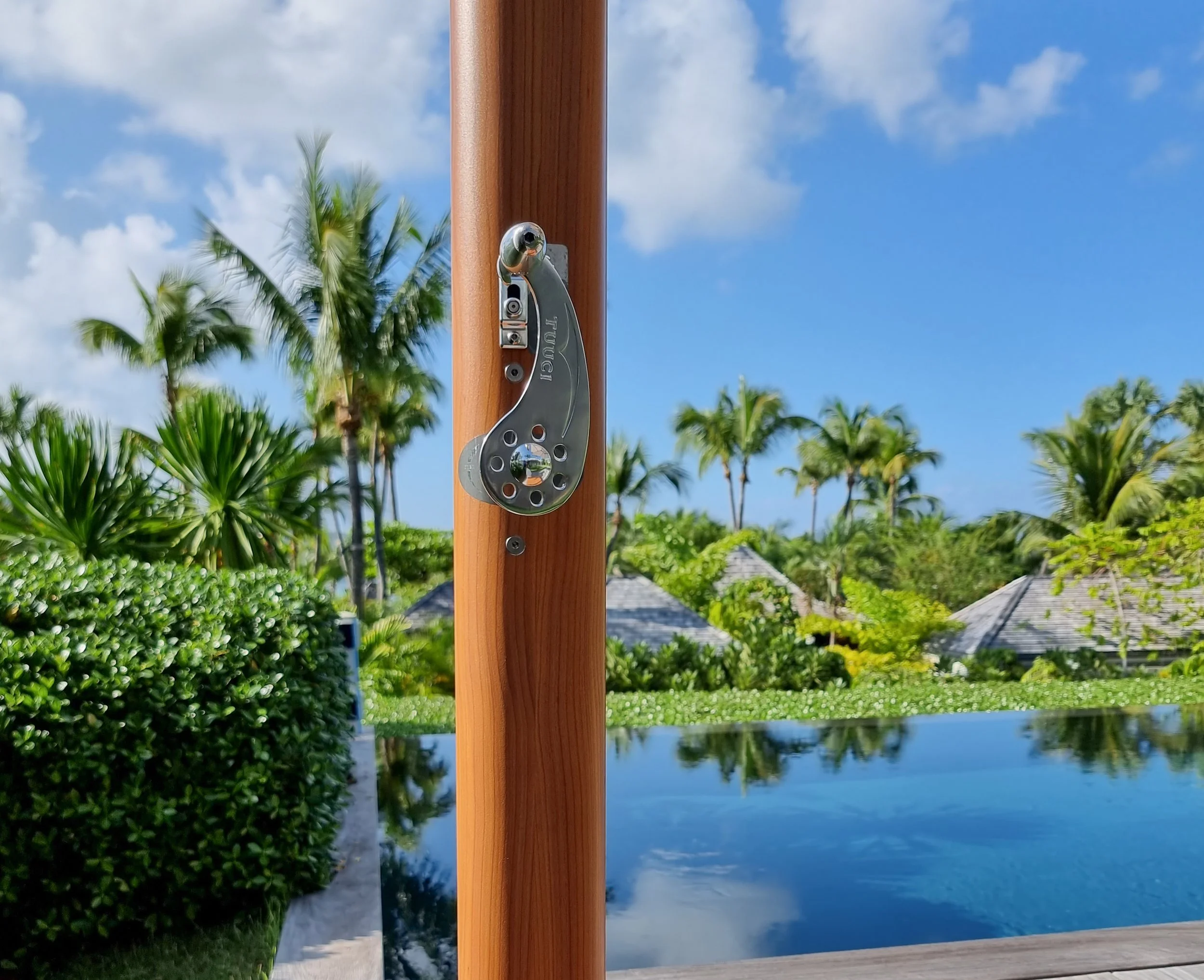 Close-up of a pulley and wooden pole with a scenic background of palm trees, a pool, and a partly cloudy blue sky.