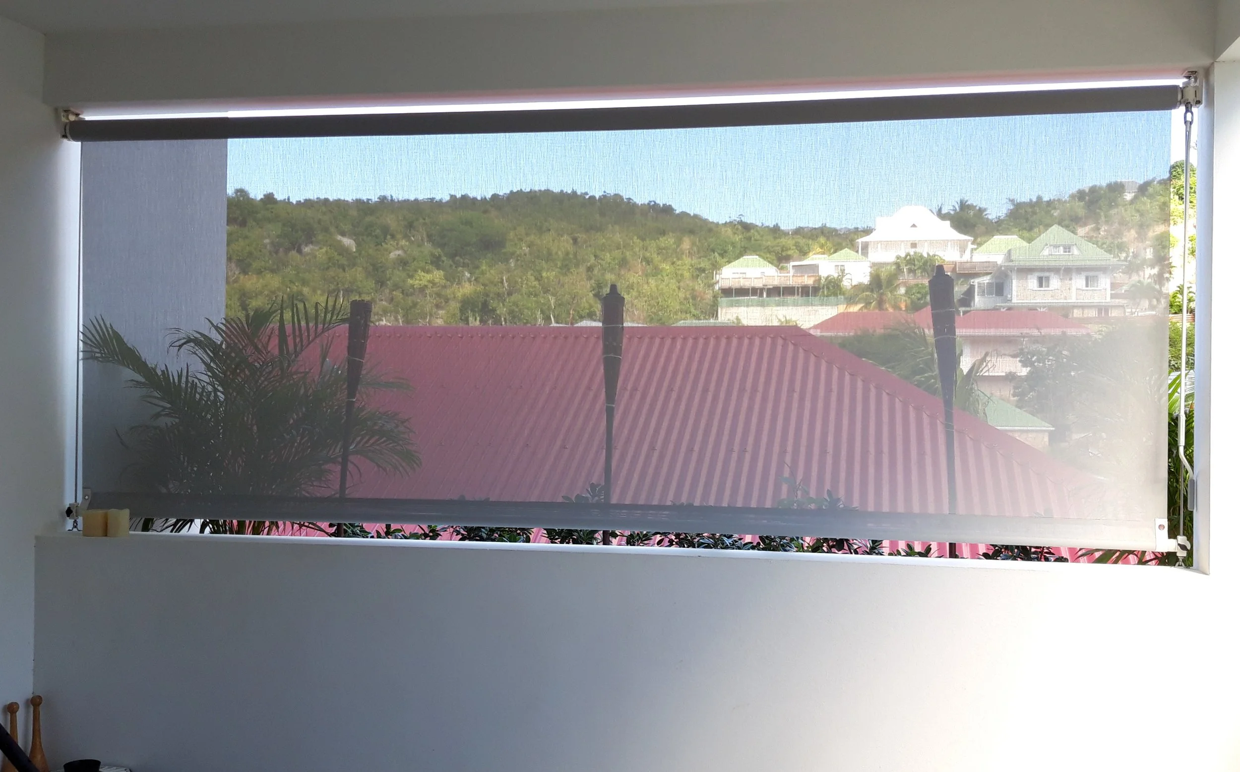 View through a rolled-up window screen showing a red roof, green hills, and houses with green roofs in the background.