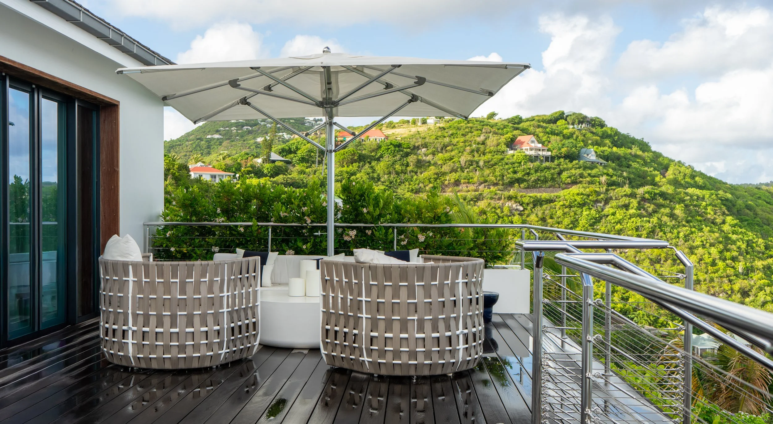 Balcony with outdoor seating, including a white cushioned sofa and chairs, under a large white umbrella, overlooking lush green hills with scattered houses and a partly cloudy sky.