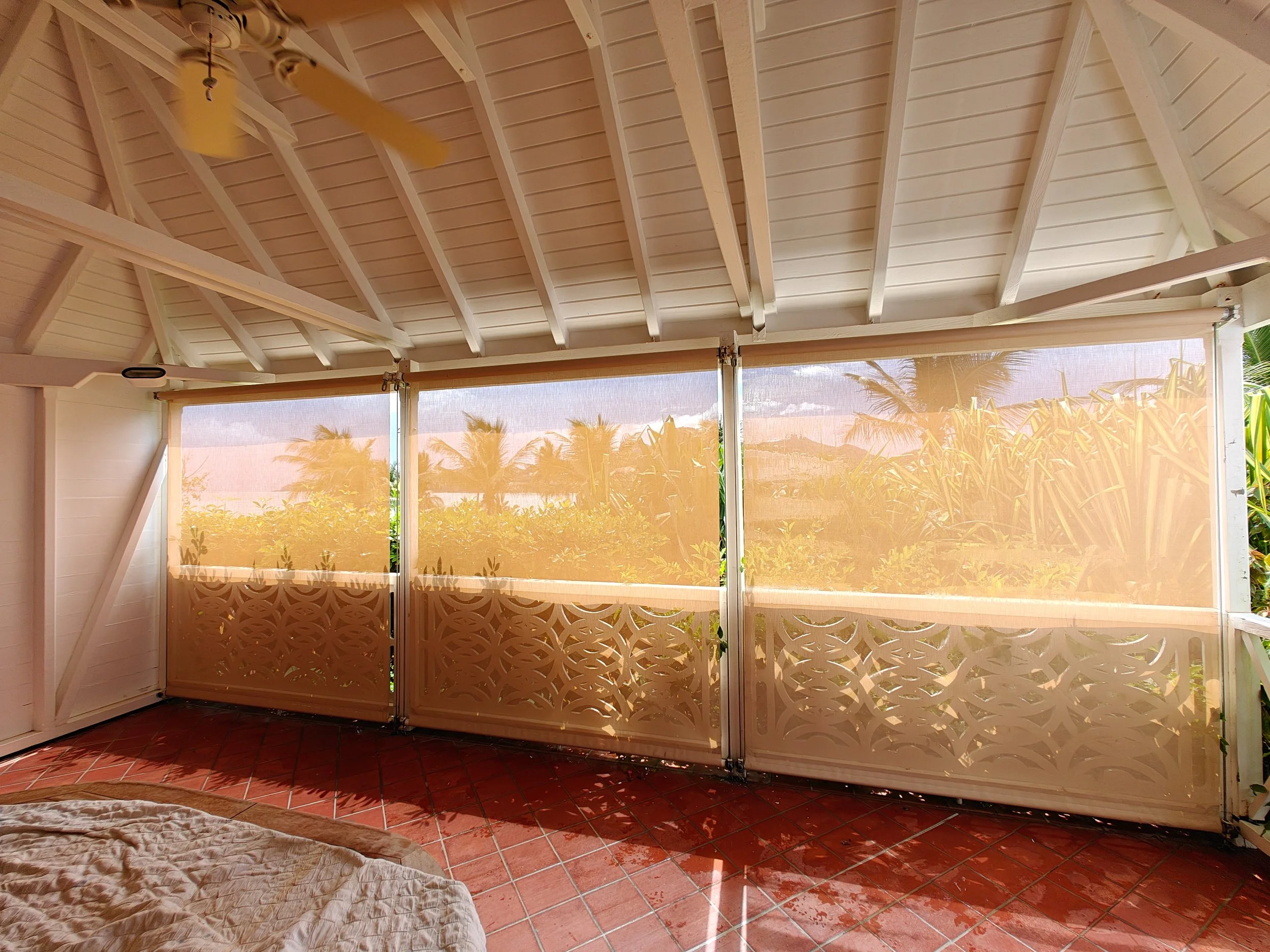 Sunlit porch area with a ceiling fan, beige decorative folding doors, and a screened opening revealing palm trees outside.
