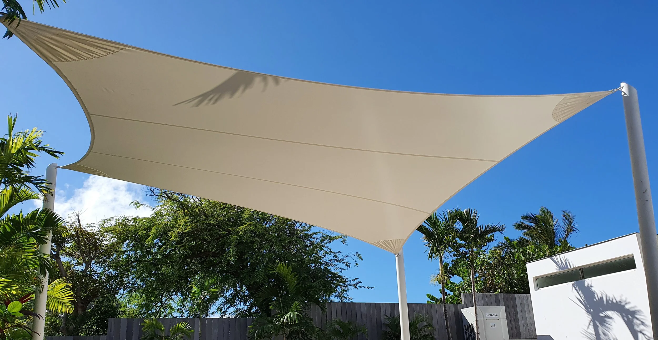 A large beige shade sail stretched between white poles outdoors, with green trees and a clear blue sky in the background.