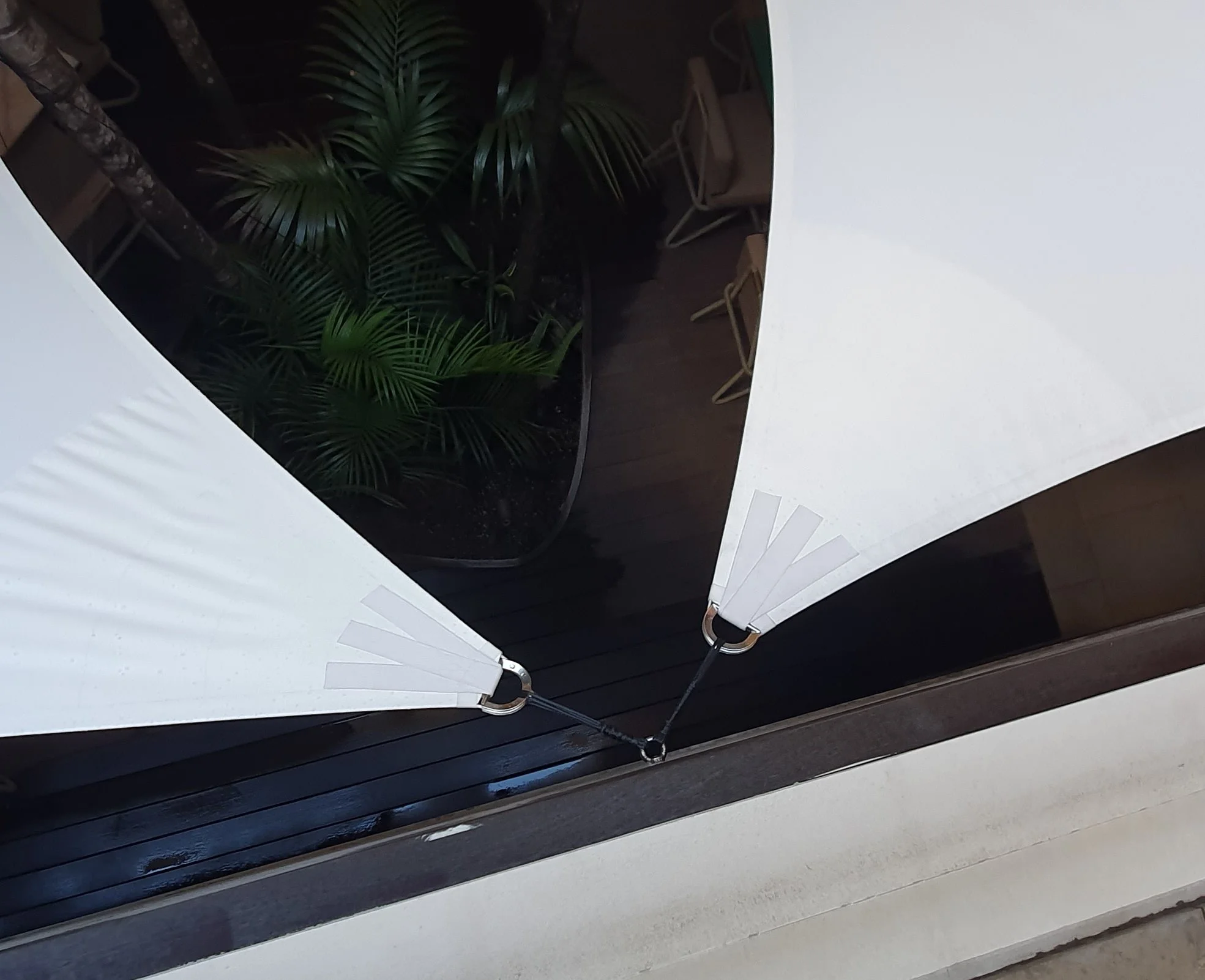The image shows a white folding fan partially open, revealing a black surface underneath. Behind the fan, there are green plant leaves and some beige chairs with metal legs.