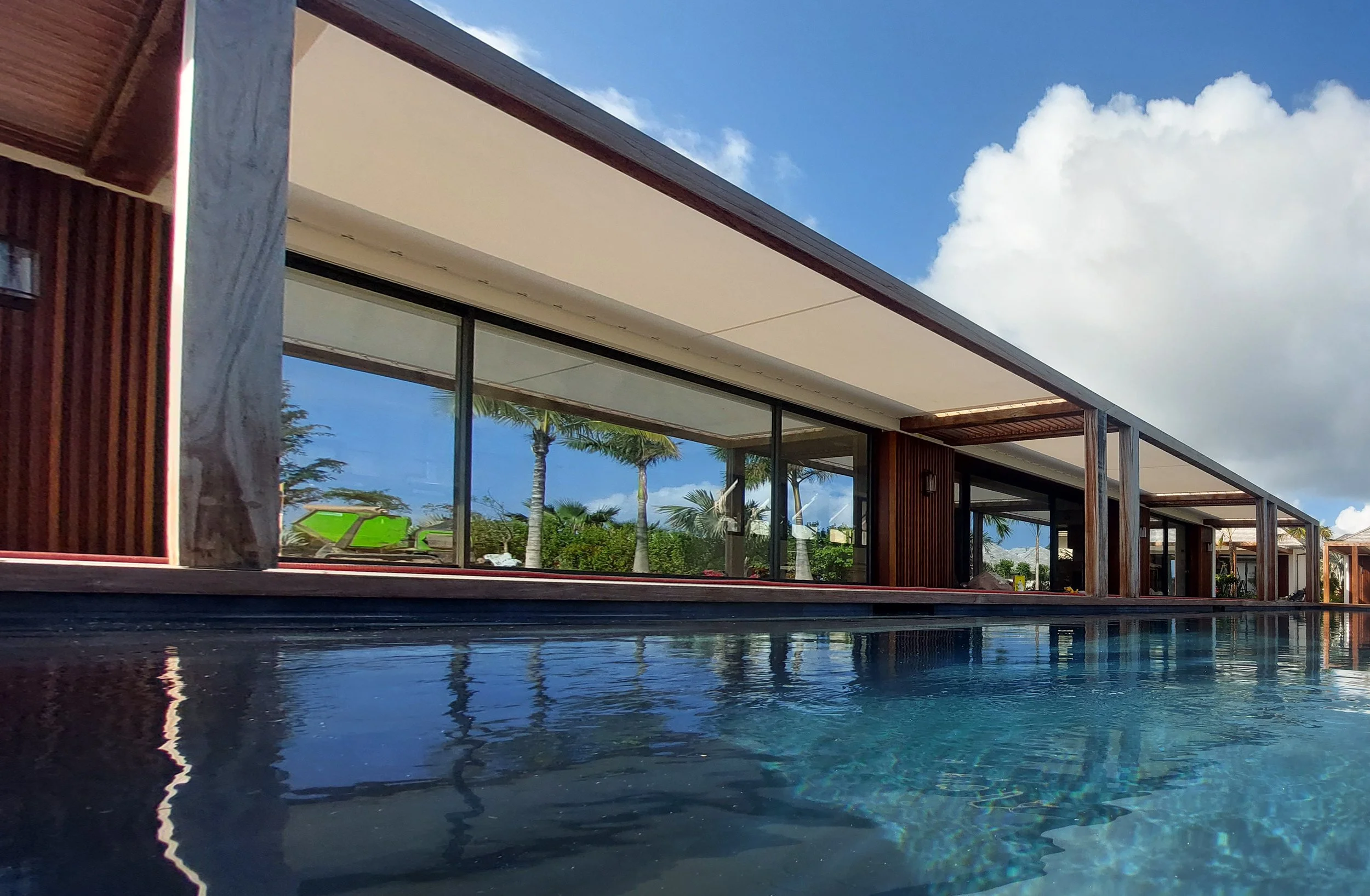 Modern house with large glass windows and a wooden exterior, reflecting palm trees and a blue sky with clouds, with a nearby swimming pool.