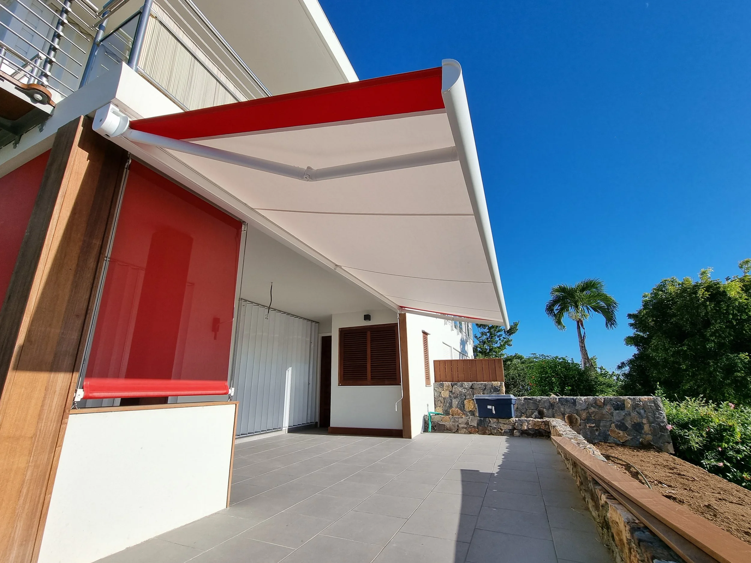 An outdoor patio area with a covered space featuring a red and white awning, a wooden fence, a palm tree, and a clear blue sky.