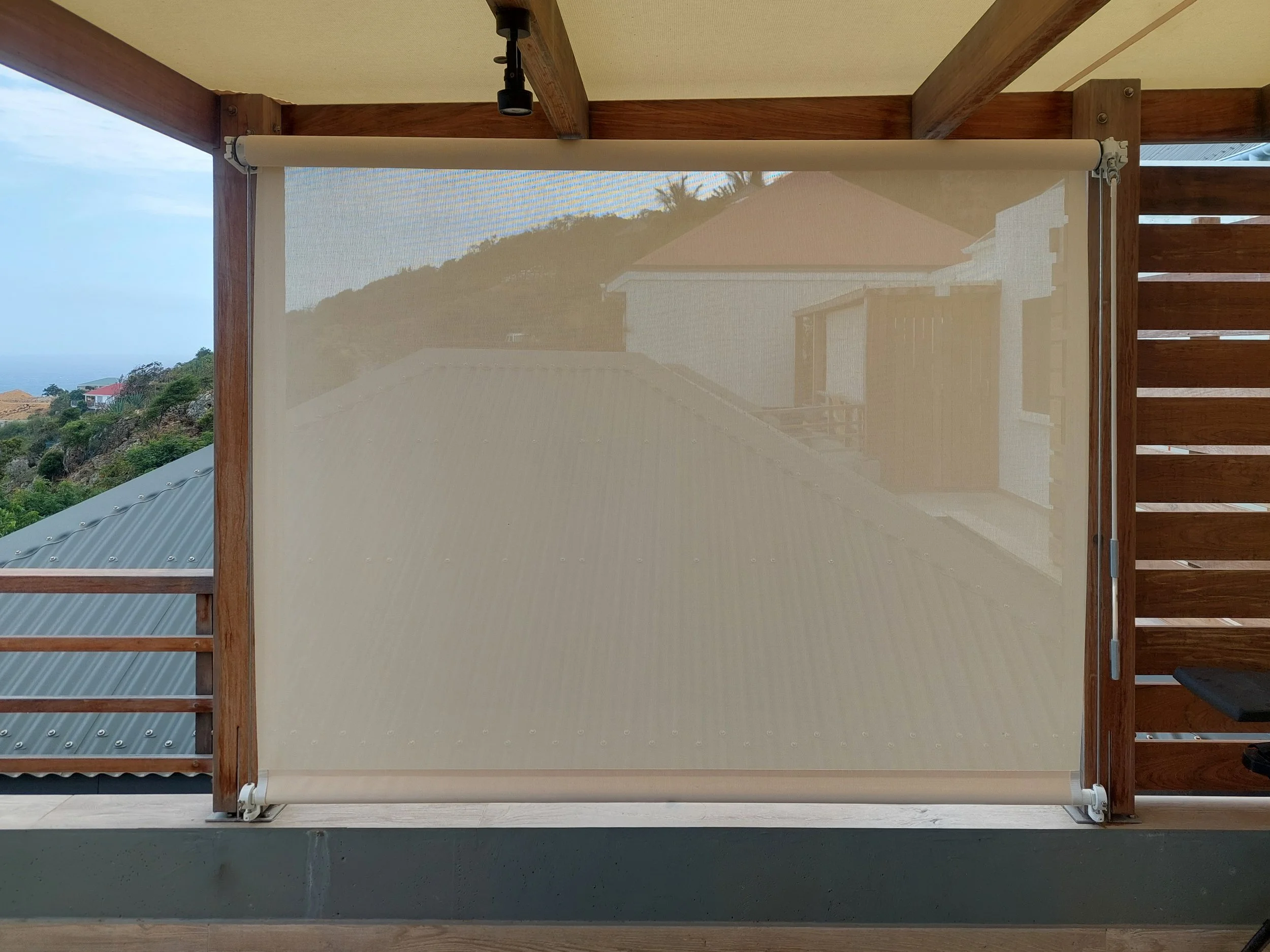 A beige roller shade partially covering a sliding glass door frame with wooden beams on the ceiling, overlooking a view of houses, greenery, and hills in the distance.