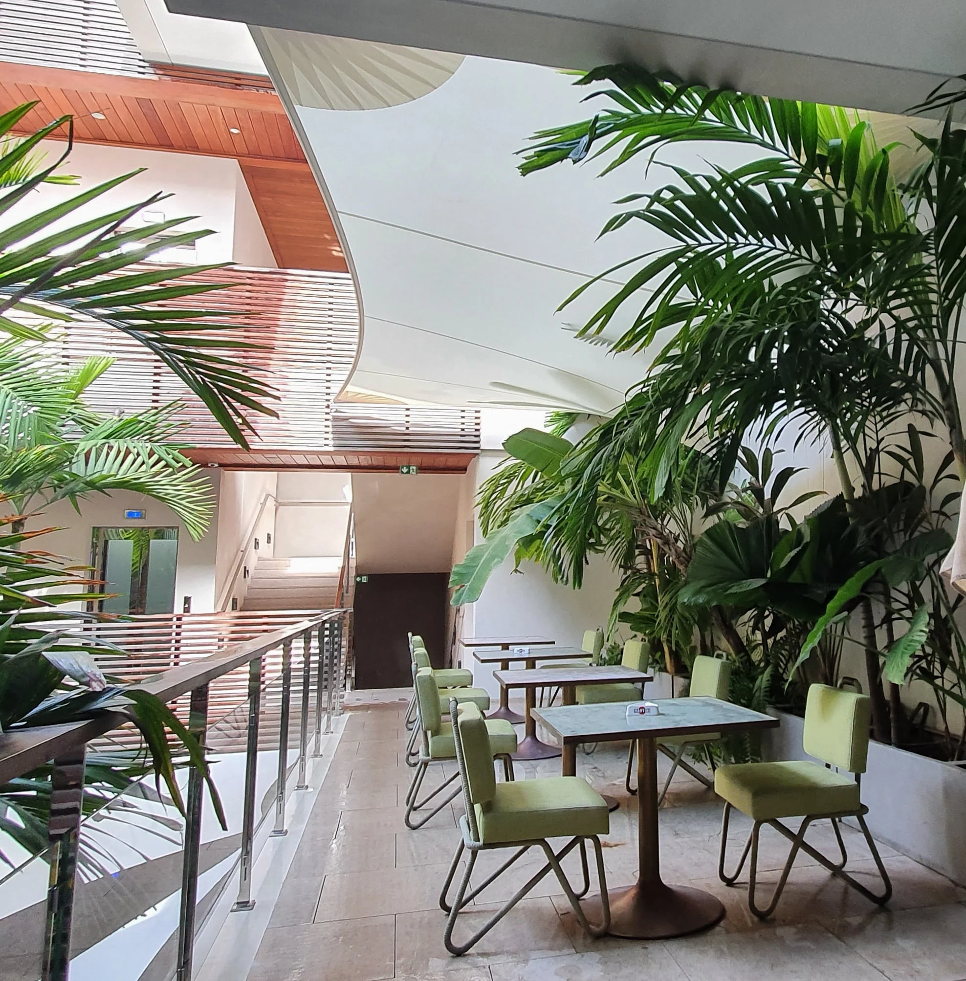 Indoor space with green chairs arranged around small tables, lush green potted plants, and modern architectural design with wooden and glass elements.