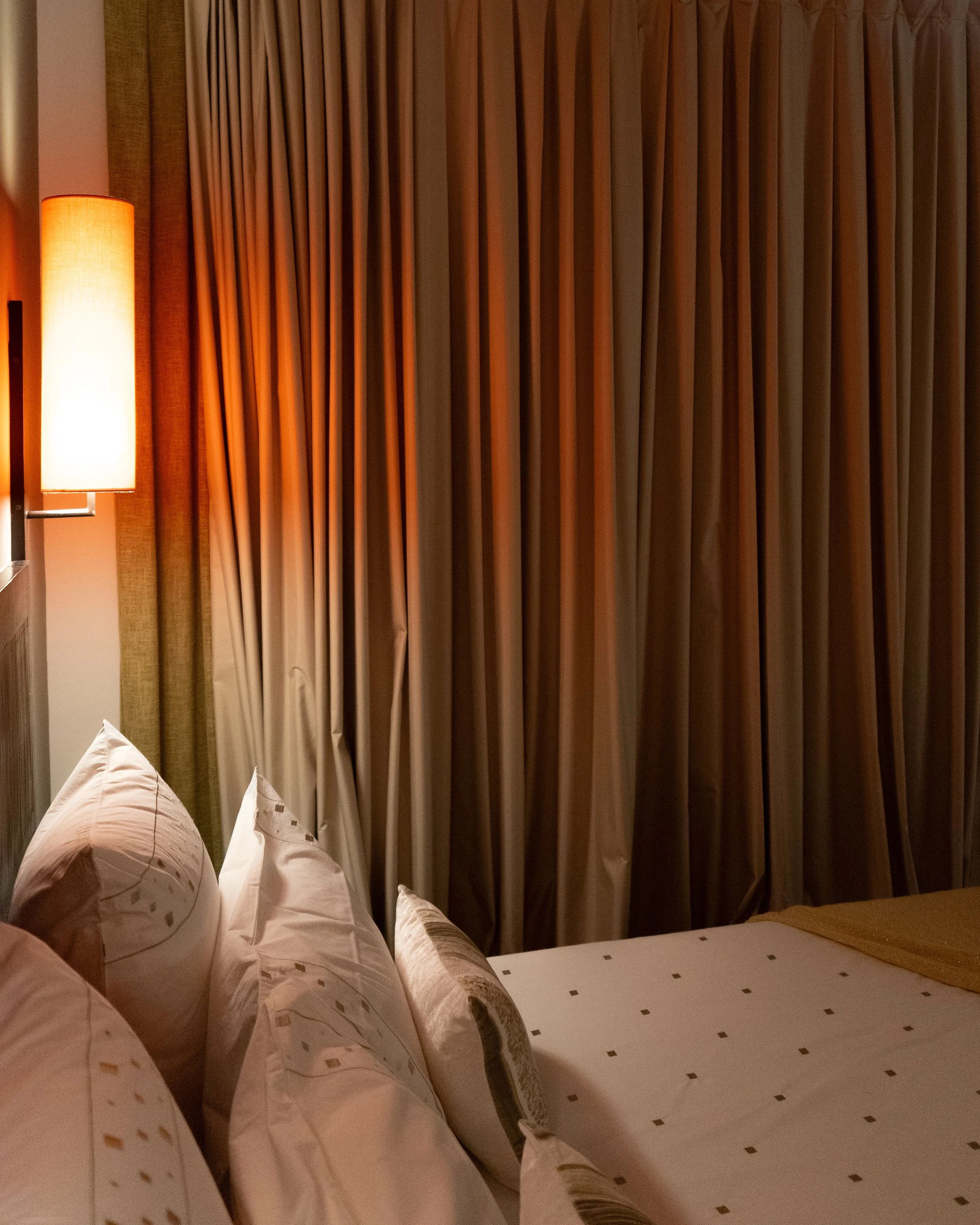 Une chambre avec des rideaux beiges, une applique murale et un lit garni d’oreillers et d’une couverture.