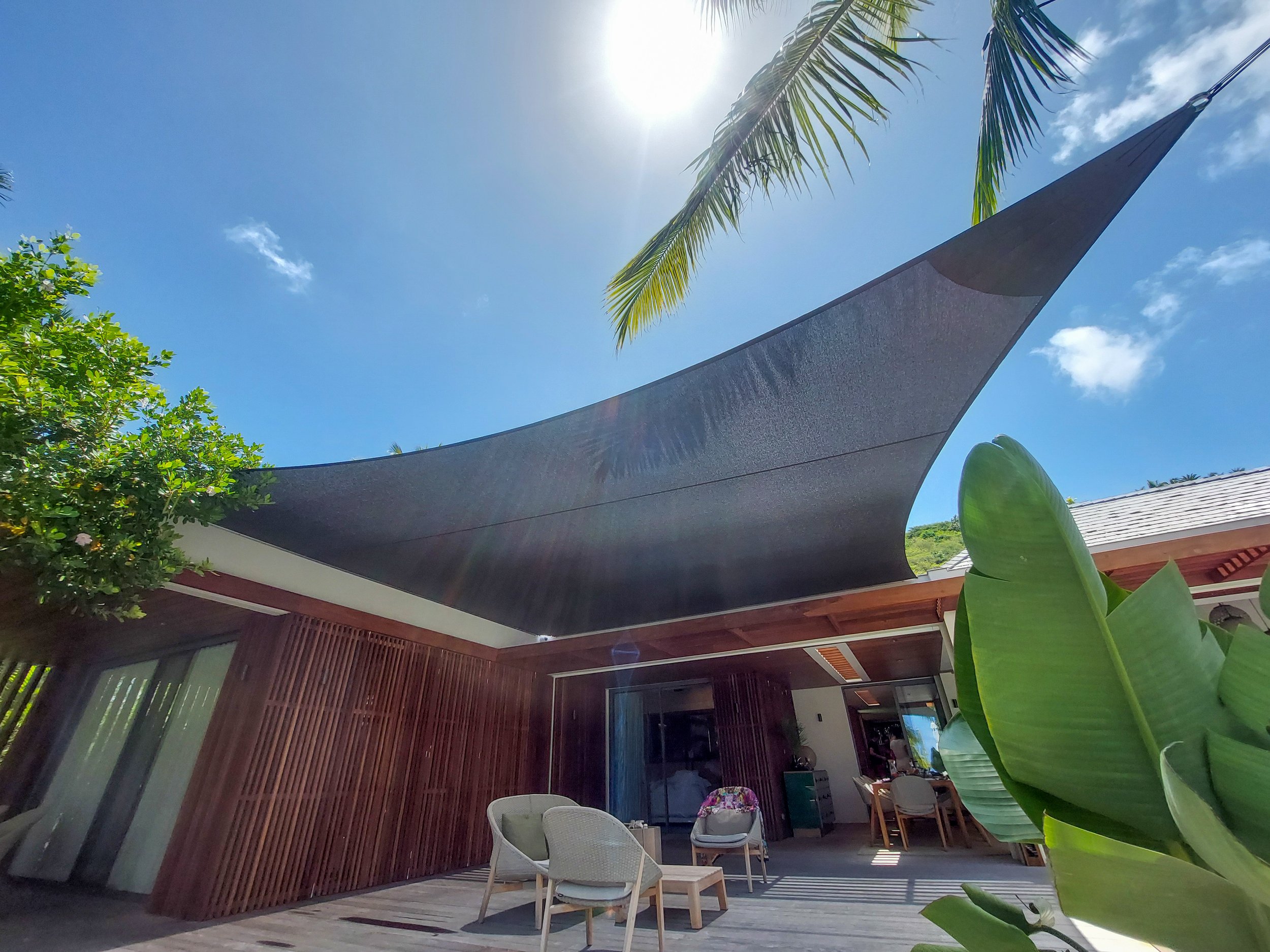 Un patio extérieur ensoleillé avec du mobilier moderne, de grandes plantes vertes et une toile d’ombrage au-dessus, sous un ciel bleu éclatant et avec des feuilles de palmier visibles.
