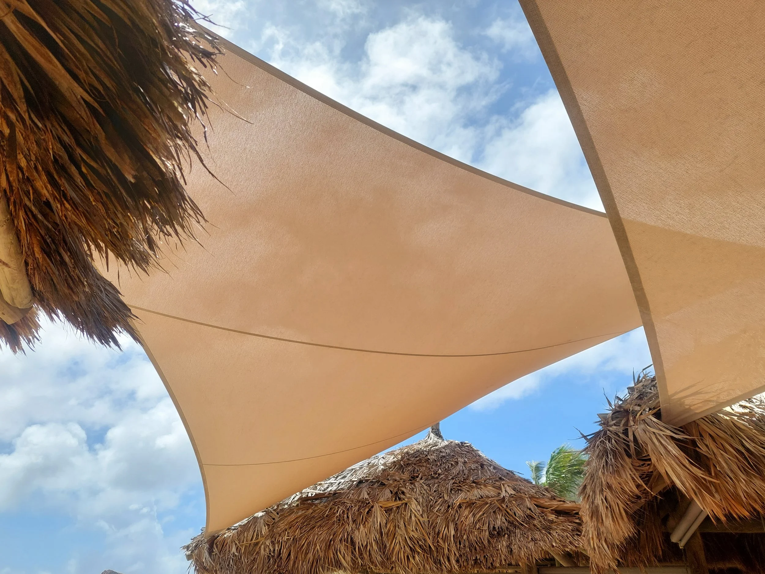 Photo taken from underneath a courtyard parasol with a thatched roof and beige canopy, with blue sky and clouds visible above.