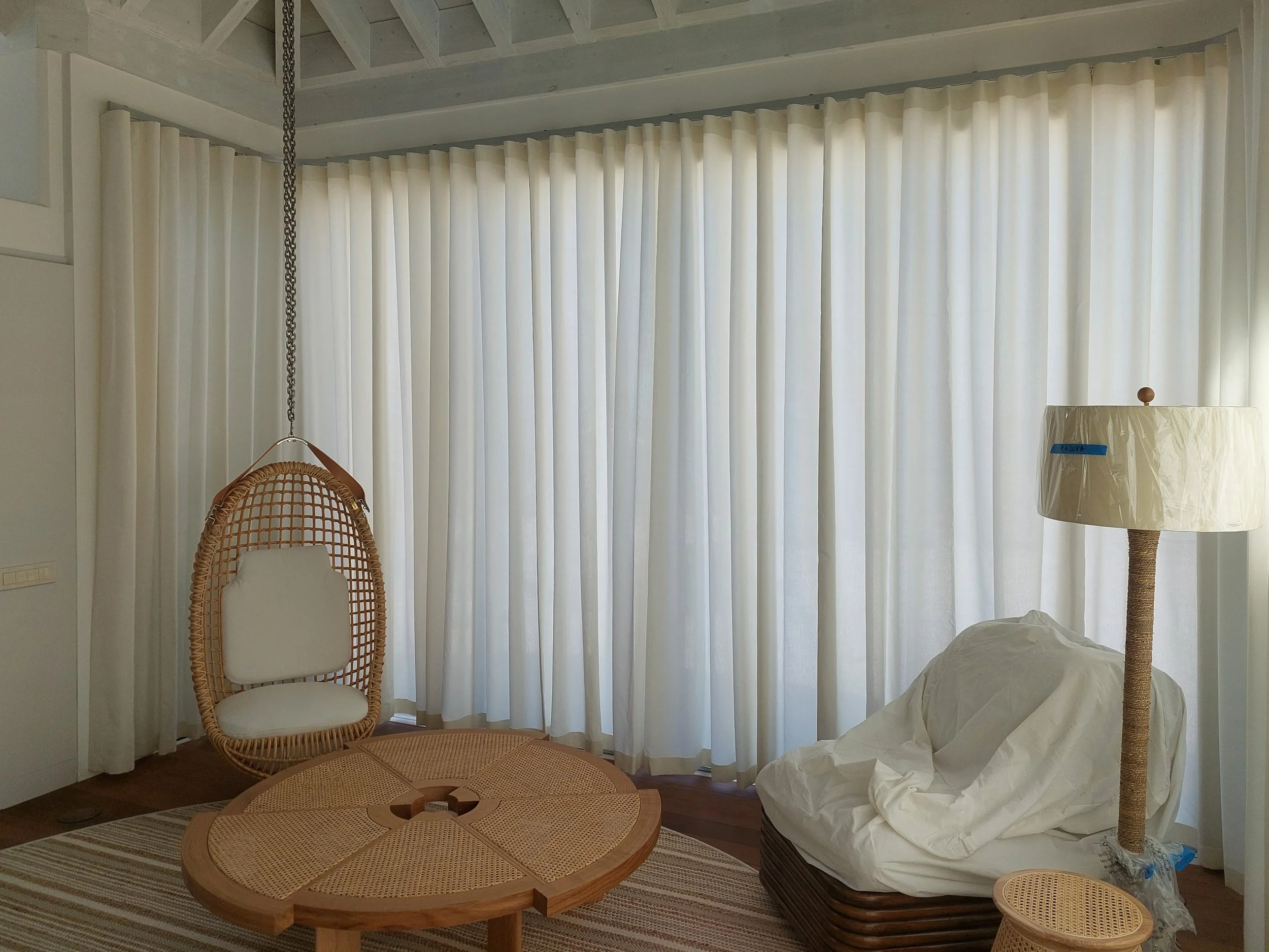 A room with cream-colored curtains covering large windows. There is a rattan hanging chair with a white cushion on the left, a round wooden coffee table in the center, and a table lamp with a fabric shade on the right, with a cloth or wrinkled fabric