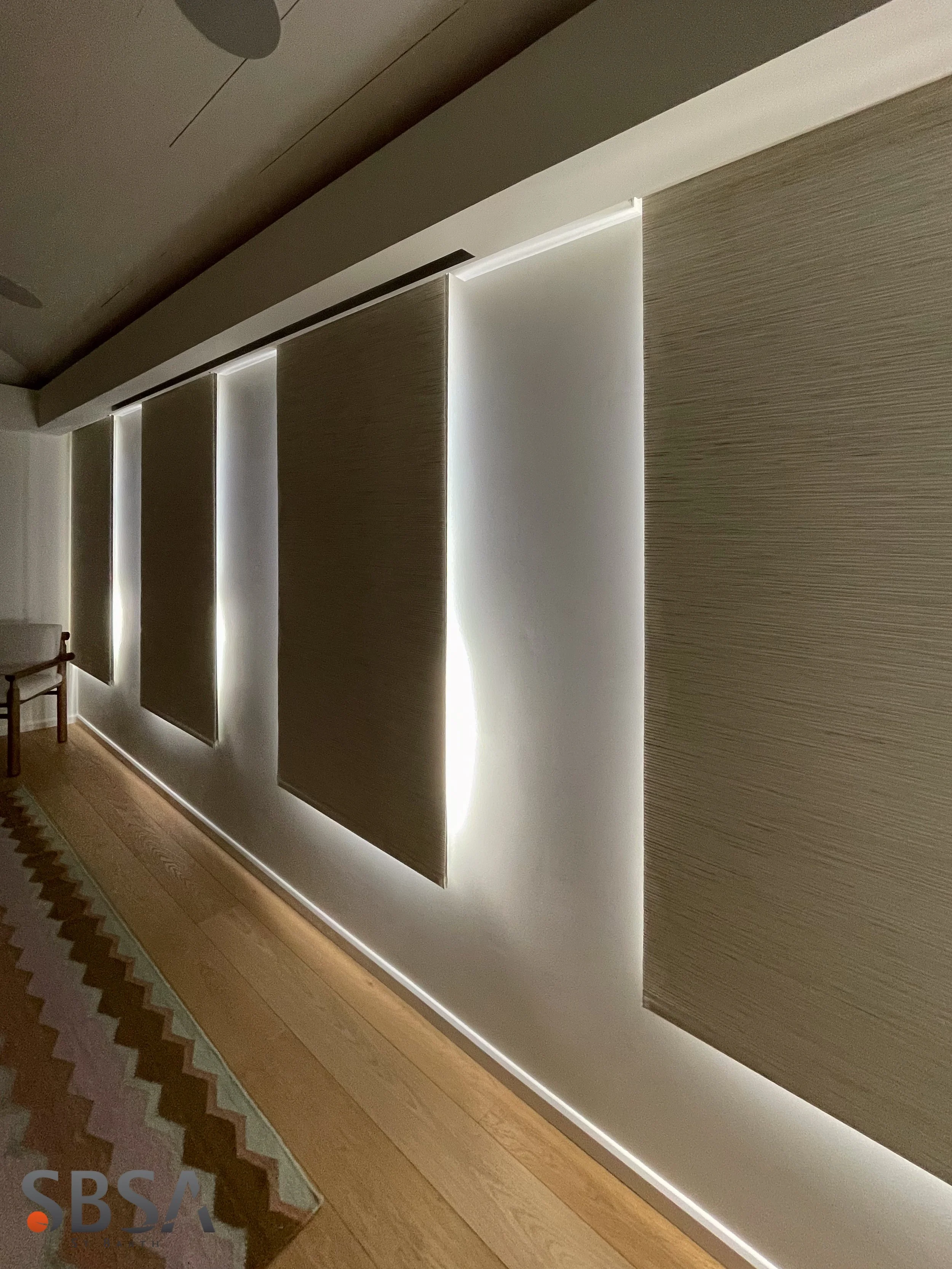 Interior view of a room with several vertical rectangular wooden paneling pieces with backlighting, a patterned rug on a wooden floor, and part of a hanging ceiling fixture visible.
