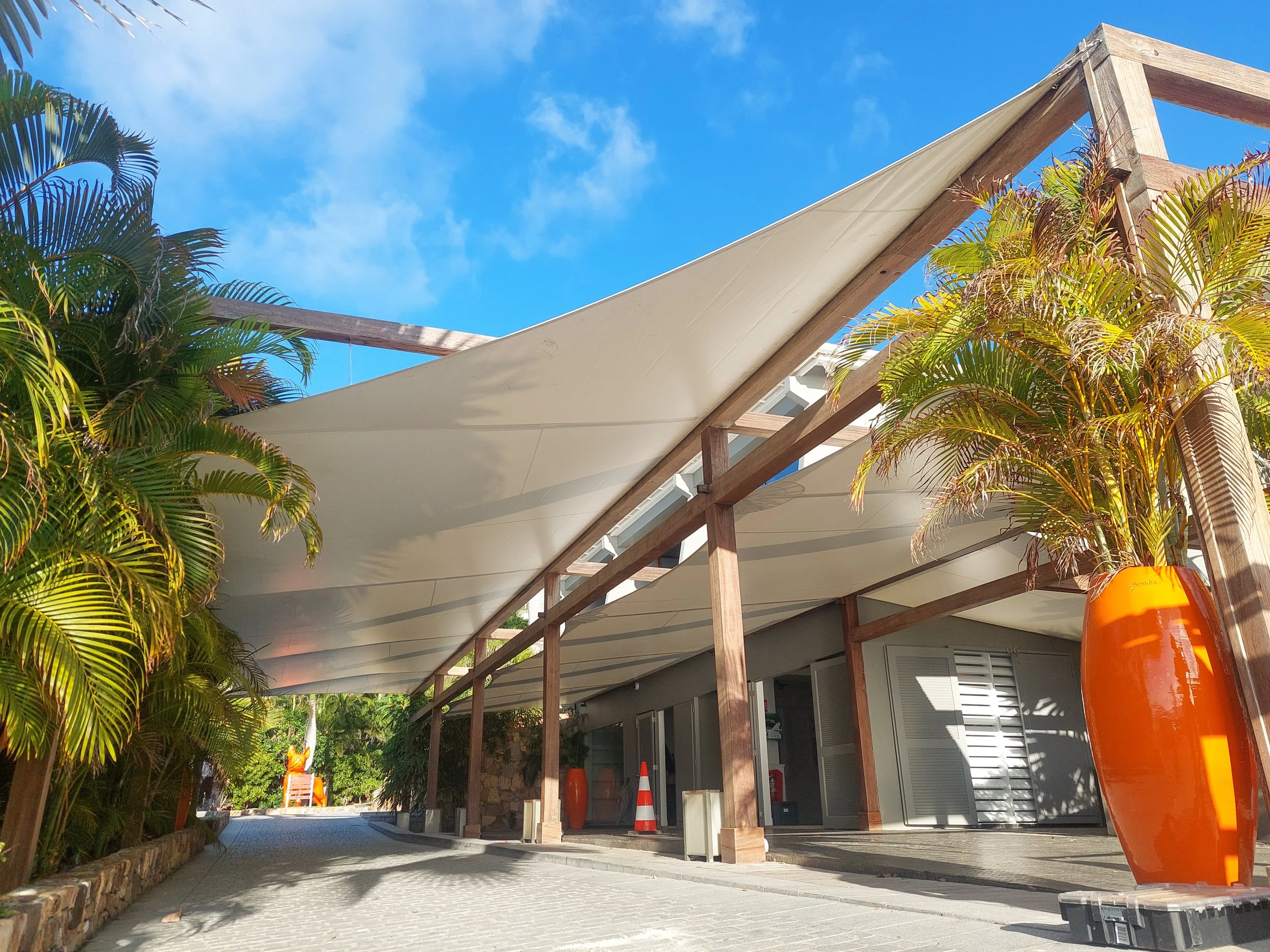 A structure with a beige shade sail canopy supported by wooden beams, with tropical palm trees on the side, orange safety cones, and a blue sky with some clouds.