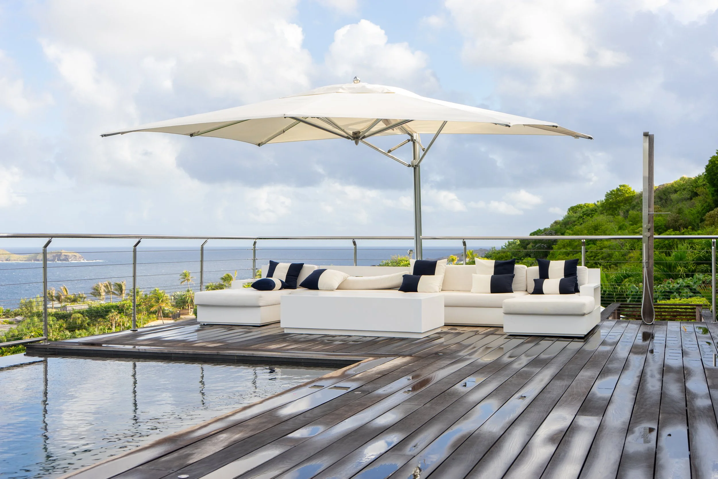 Outdoor patio with white cushioned seating, a large white patio umbrella, and a body of water in the background. The patio is made of dark wood flooring, with hills and greenery in the distance under partly cloudy skies.