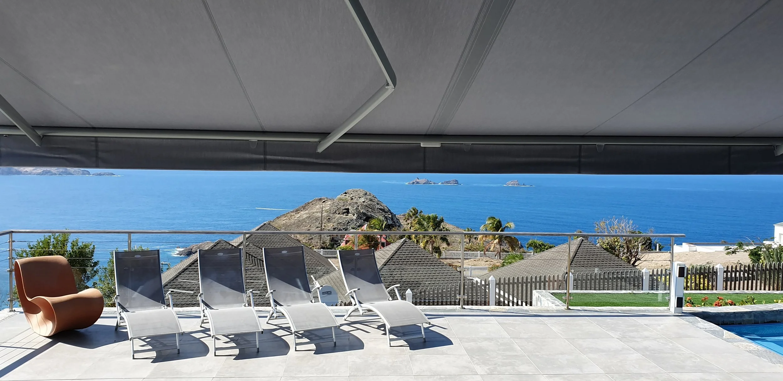 Balcony overlooking the ocean with lounge chairs, a store with arms, and a safety railing, showing rocky islands and water in the distance.