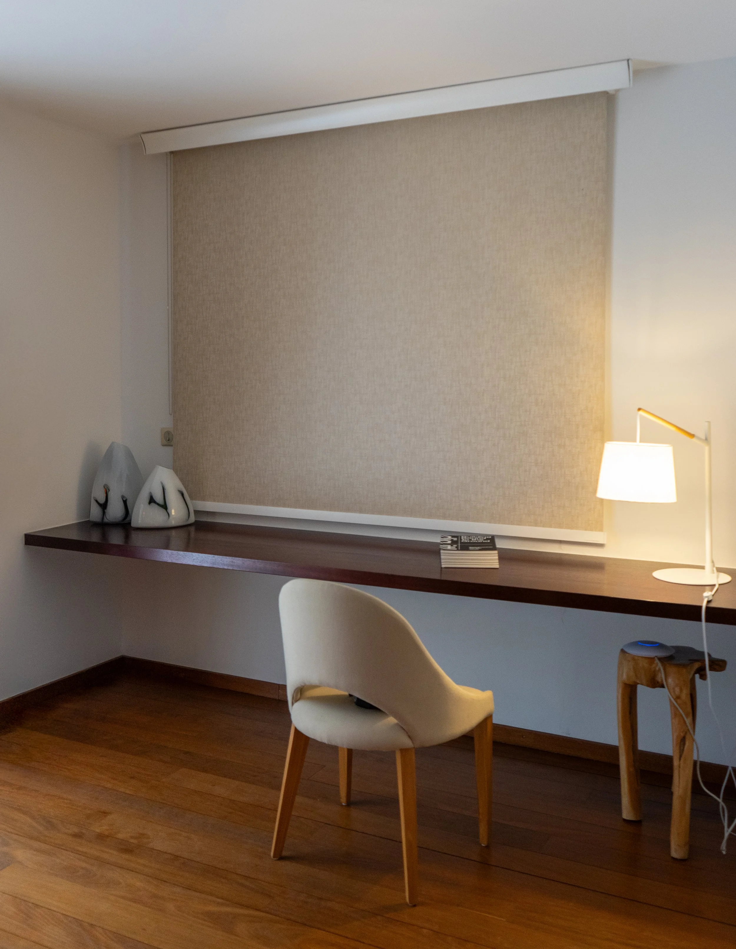 Un coin chaleureux de la pièce comprenant un petit bureau en bois, une chaise blanche rembourrée, une lampe de table et des figurines décoratives en céramique représentant des animaux, avec une fenêtre équipée d’un store enrouleur beige.