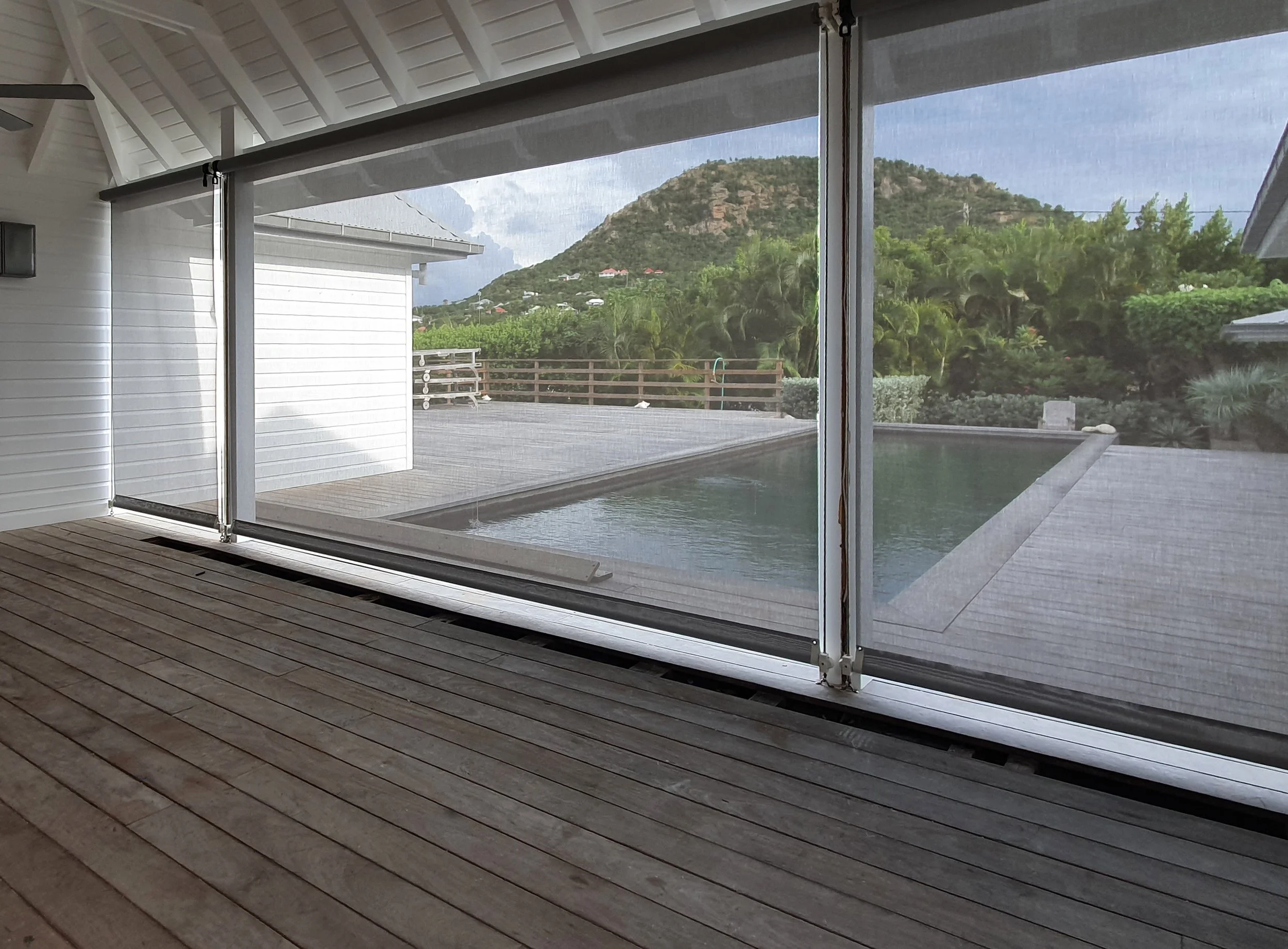 View from inside a house through a large glass sliding door showing a wooden deck, a swimming pool, greenery, and a mountain in the background.
