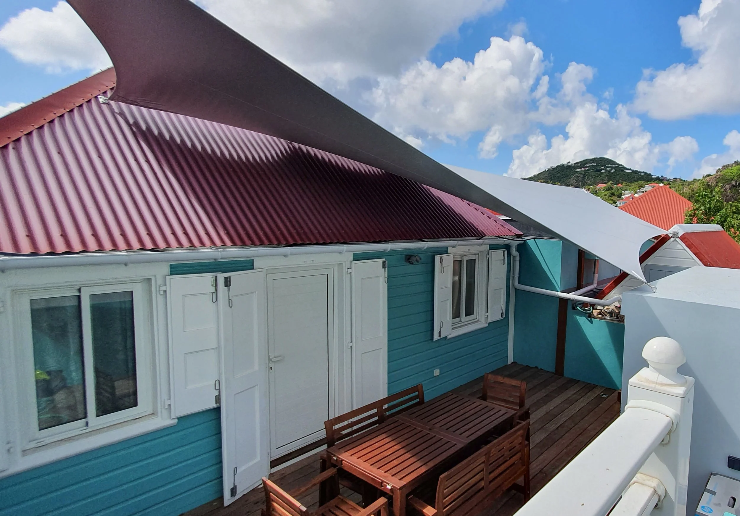 A backyard patio area with wooden table and chairs, a blue house with white doors, red roof, and a deck, under a partly cloudy sky with mountain hills in the background.