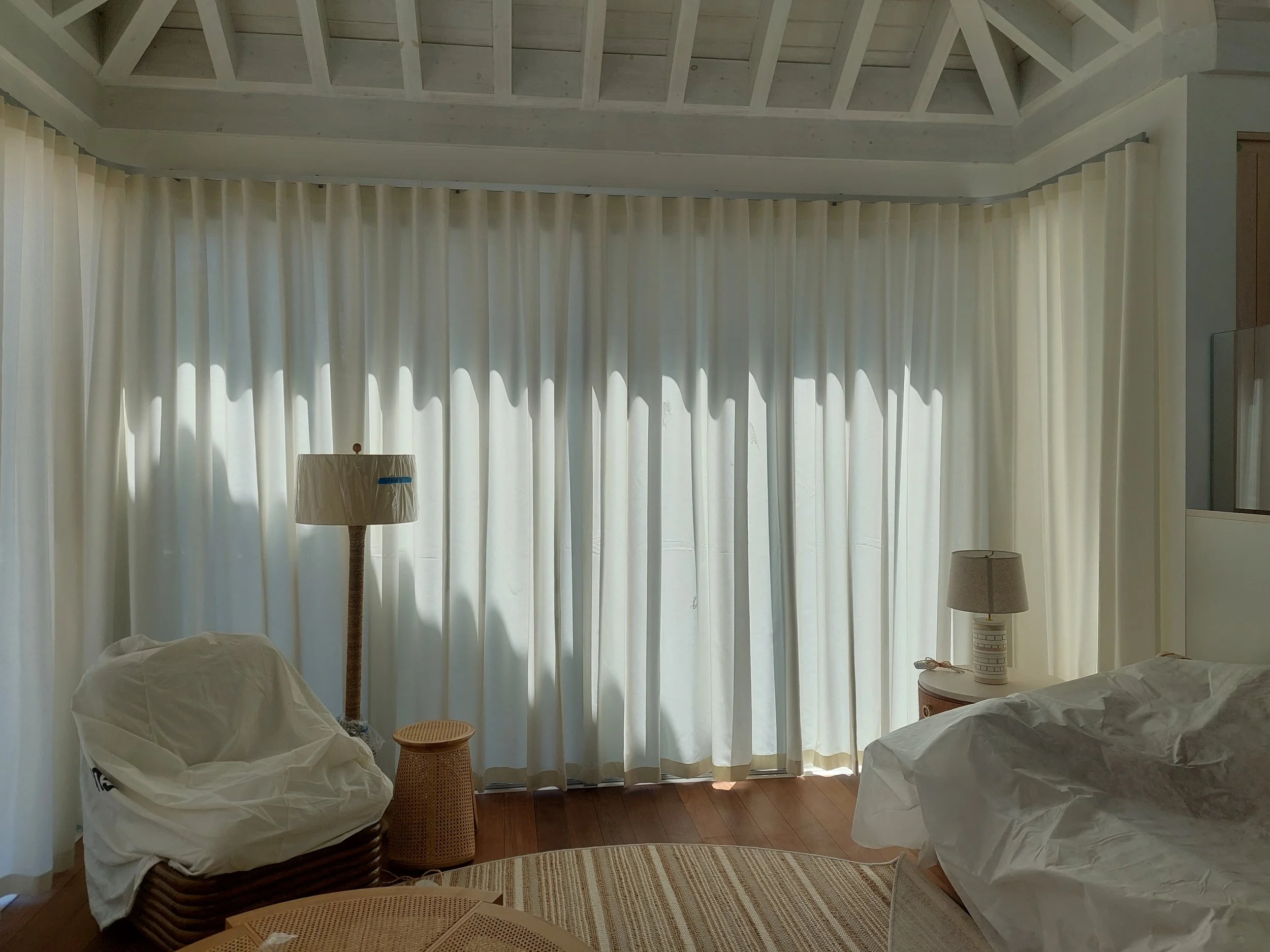 A living room with cream-colored curtains covering large windows, a lamp on a side table, and furniture wrapped in white protective covers.