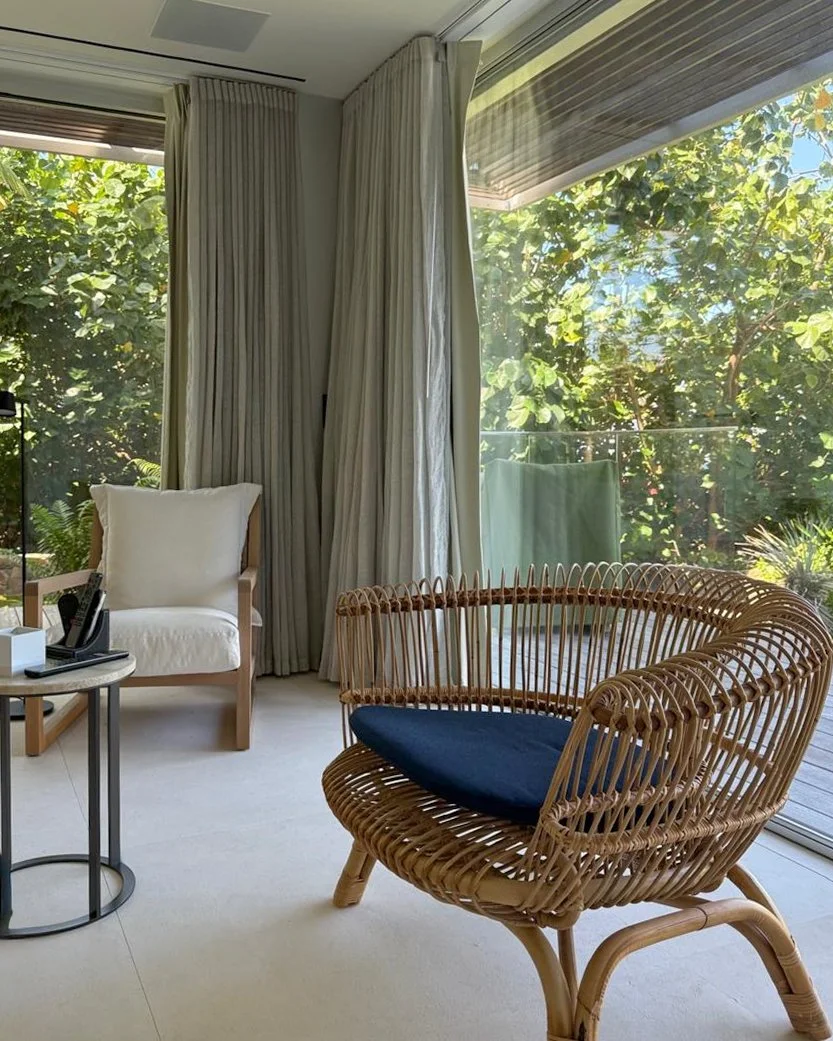 A cozy indoor living space with large windows showing green outdoor foliage. There is a white cushioned armchair, a round side table with a remote control and a small object, and a rattan chair with a black cushion. Light-colored curtains hang beside the windows.