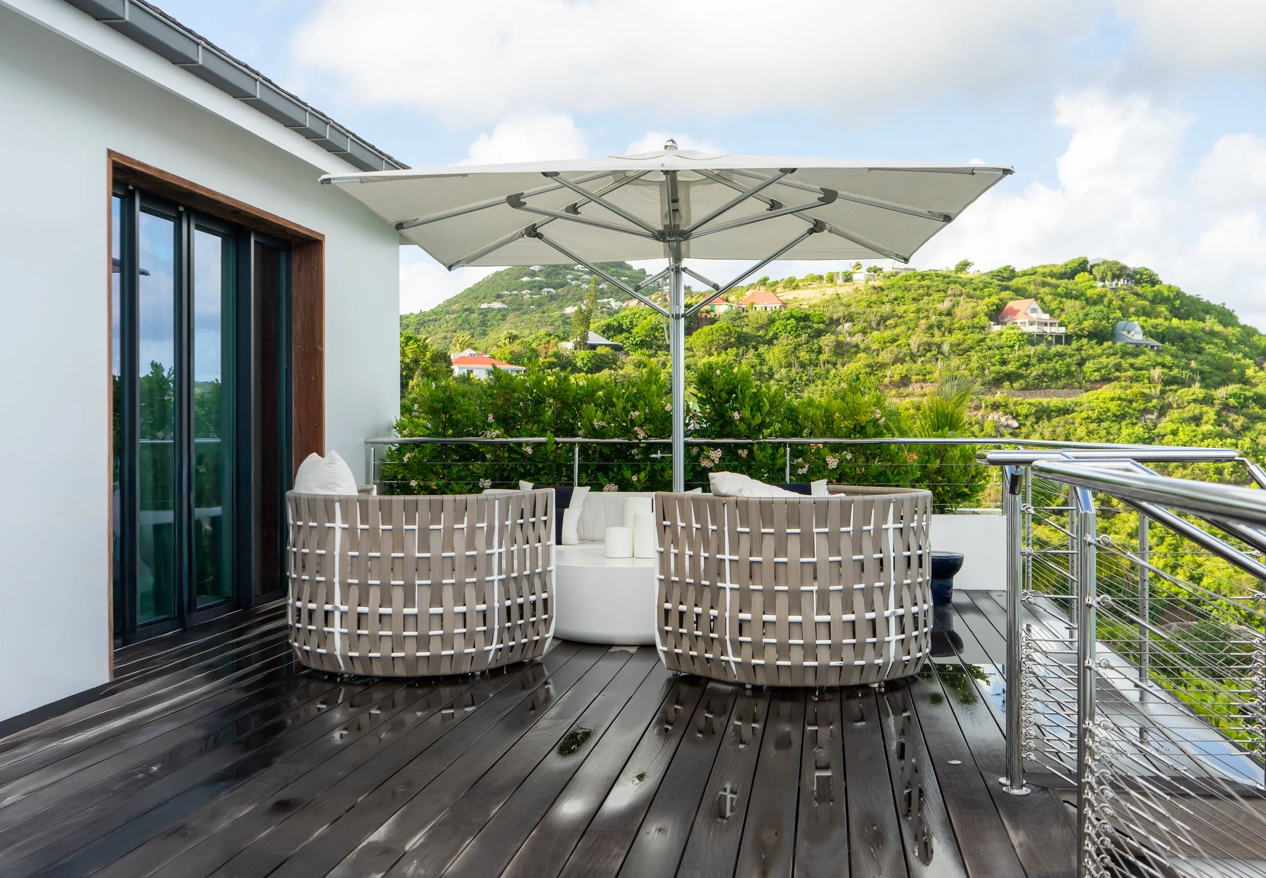 Outdoor balcony with wooden flooring, white wicker furniture, large umbrella, and lush green hill with houses in the background.