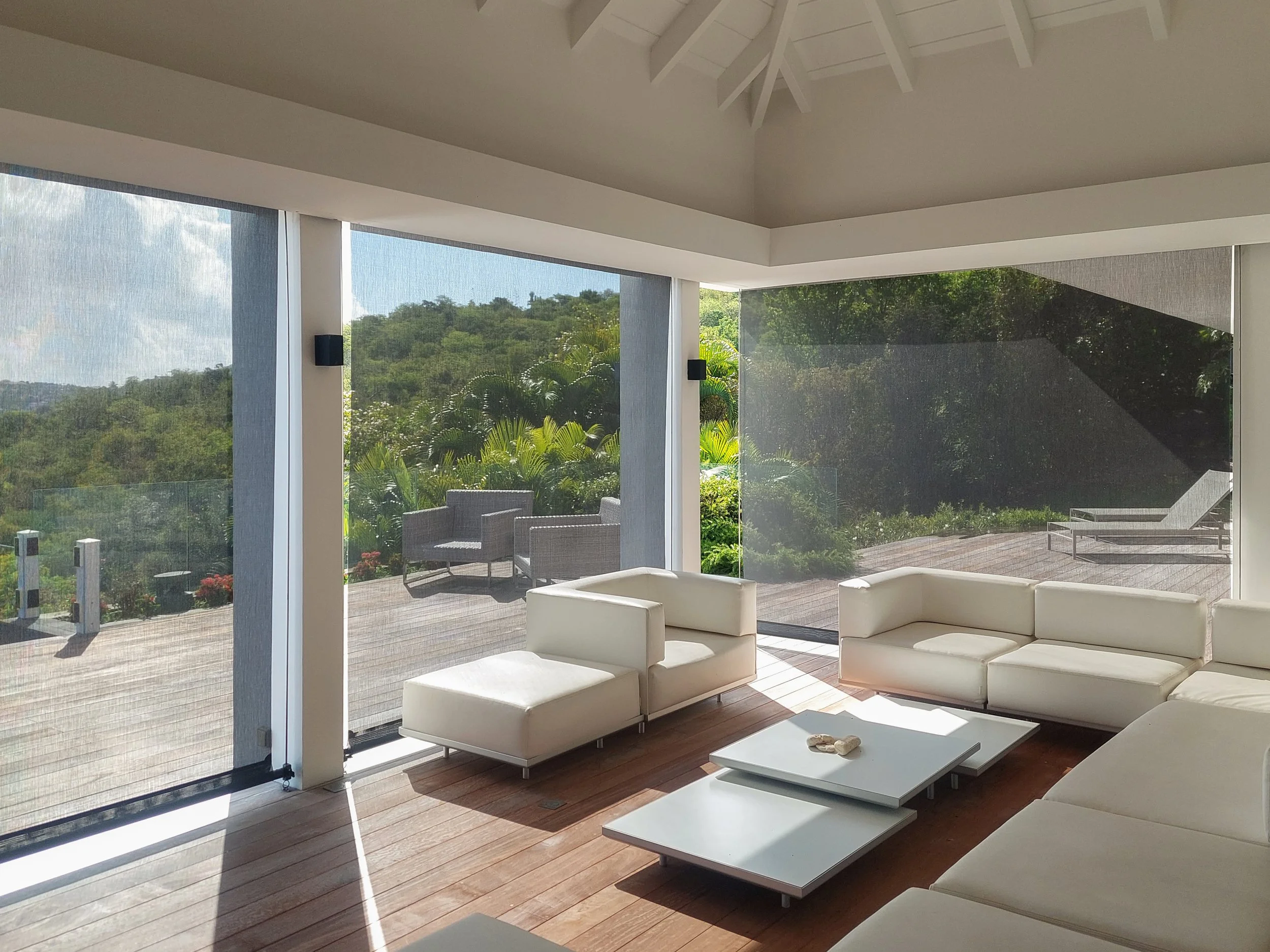 Modern living room with large glass windows showcasing green outdoor scenery, featuring white sofas and sleek white coffee tables on a wooden floor.