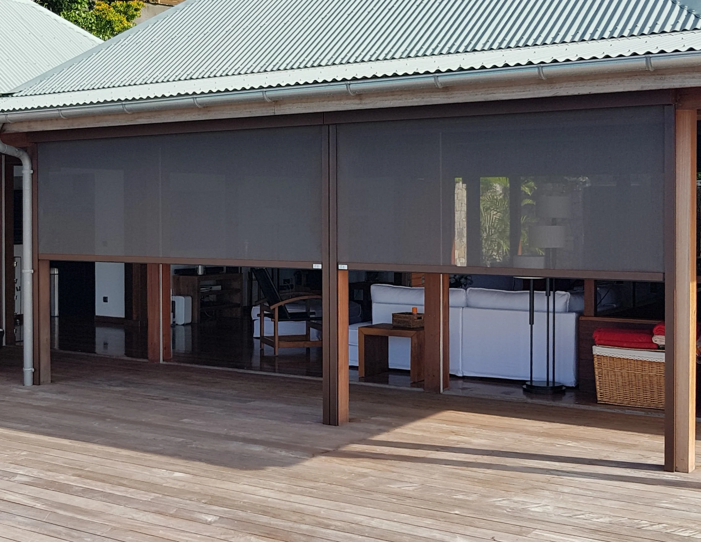 An outdoor patio with a wooden deck, a structure with a corrugated metal roof, screened-in area with furniture inside, including a white sofa, wooden chairs, a wicker basket, and a side table, surrounded by greenery.
