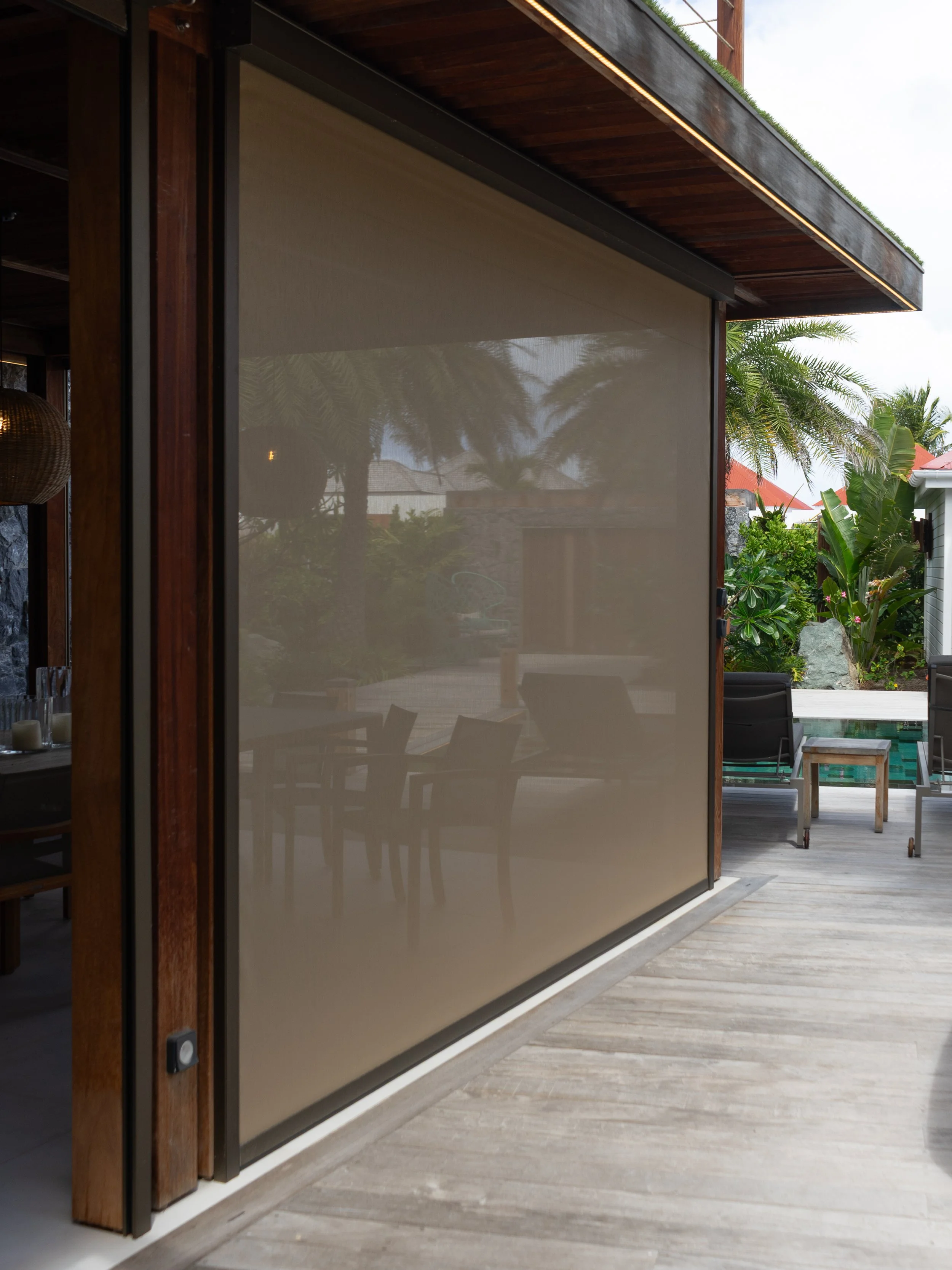 A modern outdoor patio area with wooden beams, a retractable beige fabric sunshade, lush greenery, trees, and a swimming pool in the background.