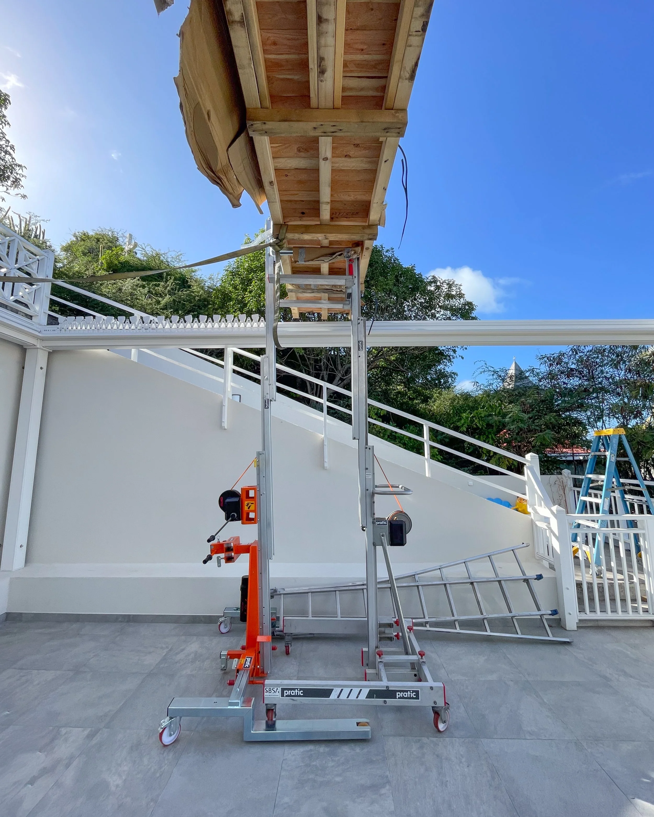 A metal scaffolding and lift platform set up on a patio with a white railing, against a background of trees, blue sky, and a white building. The electric pergola is in the process of being assembled or used, with tools attached and wires visible.