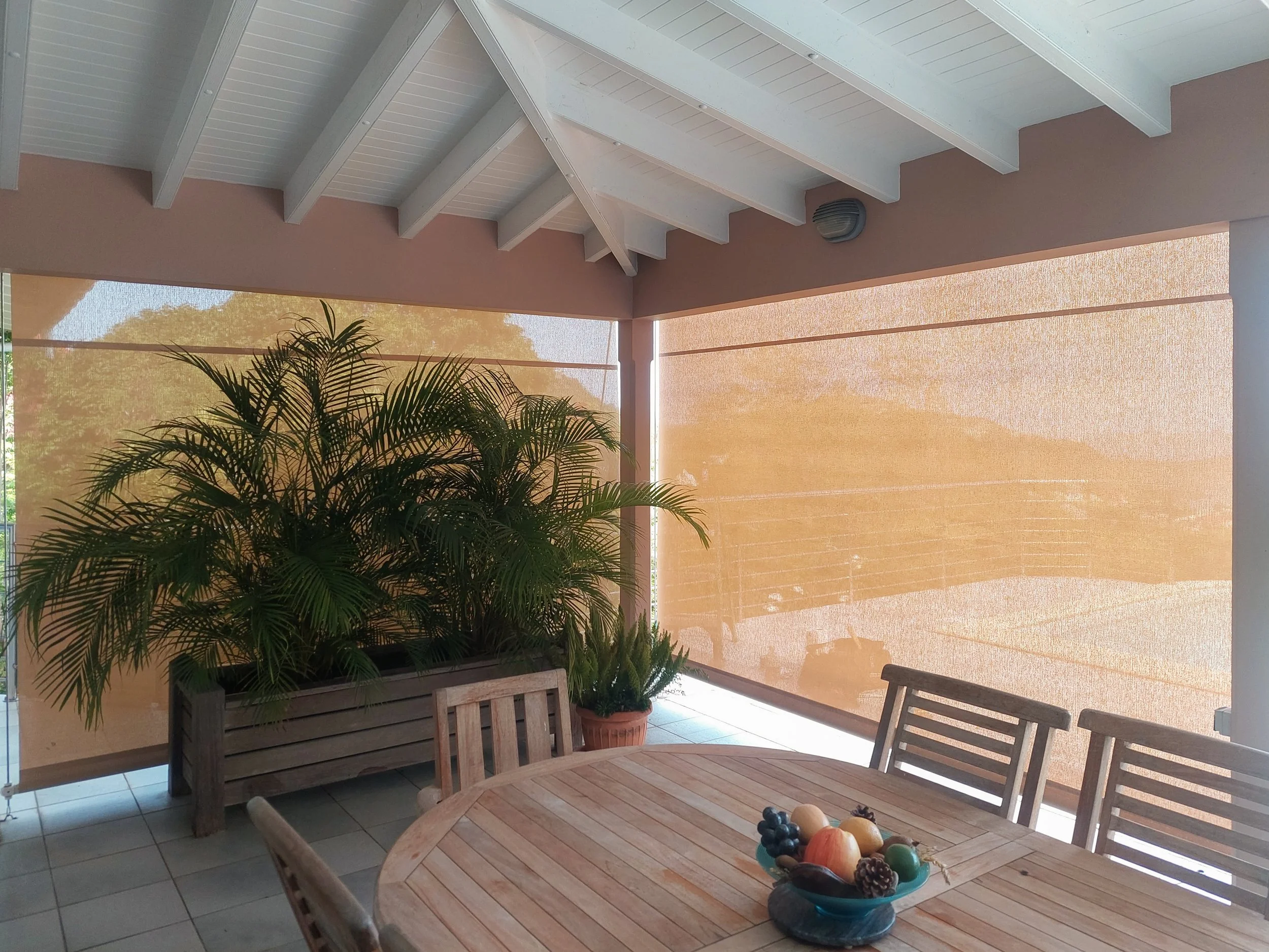 An outdoor patio area with a wooden dining table and matching chairs. There are potted plants, including a large leafy plant, and a bowl of assorted fruits on the table. The patio has a beige wall with a wooden ceiling featuring exposed beams, and a 