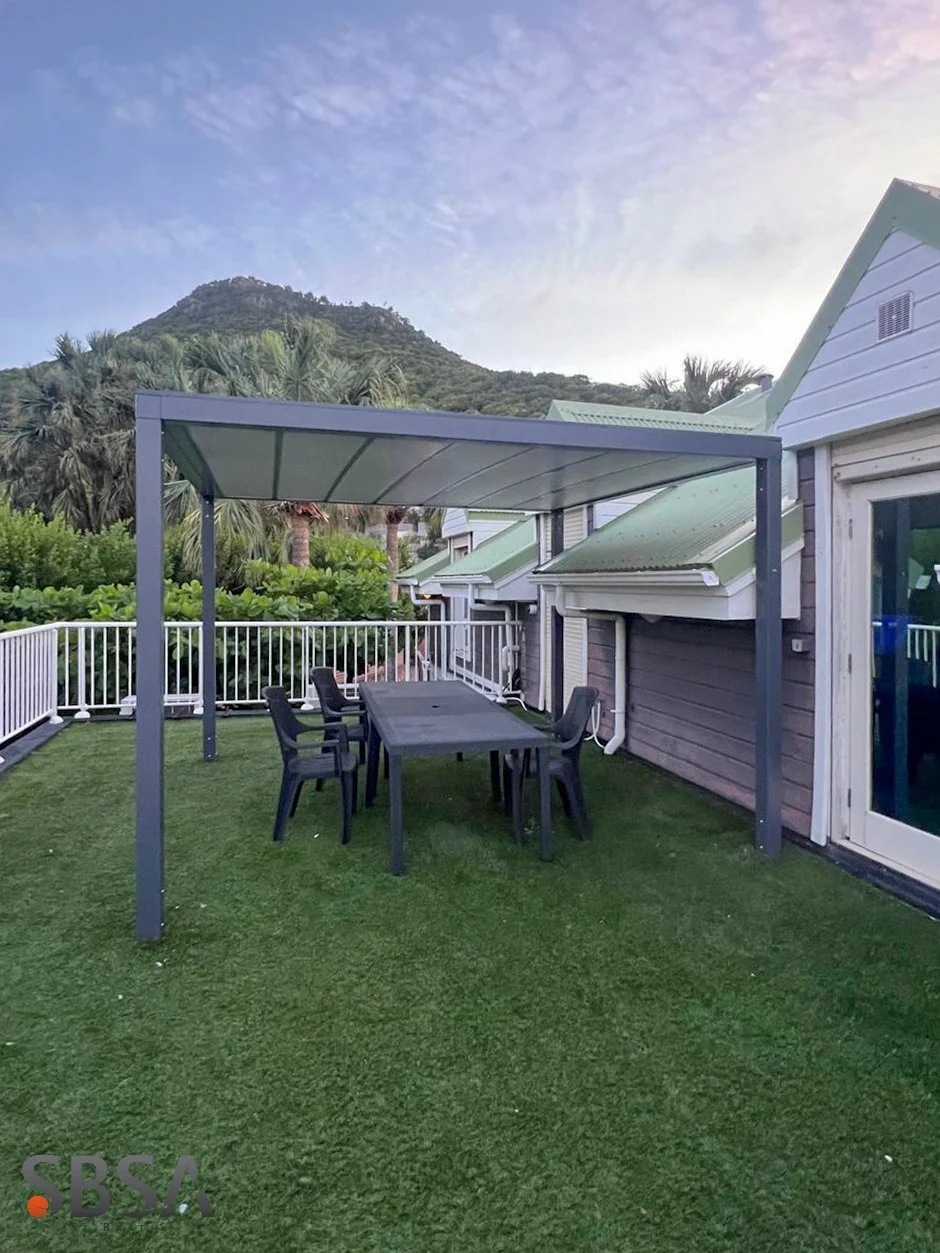 Outdoor patio with a black table, six black chairs, enclosed by a white railing, with a mountain and trees in the background, under a partly cloudy sky.