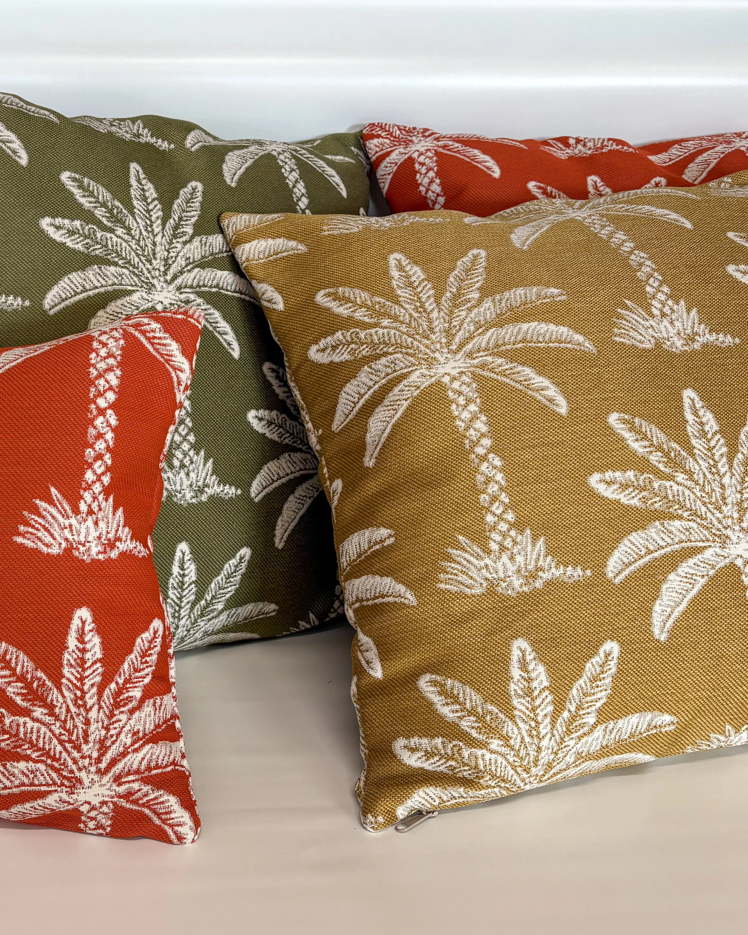 Three decorative pillows with a palm leaf pattern in different colors—green, red, and gold—placed on a white surface against a white background.