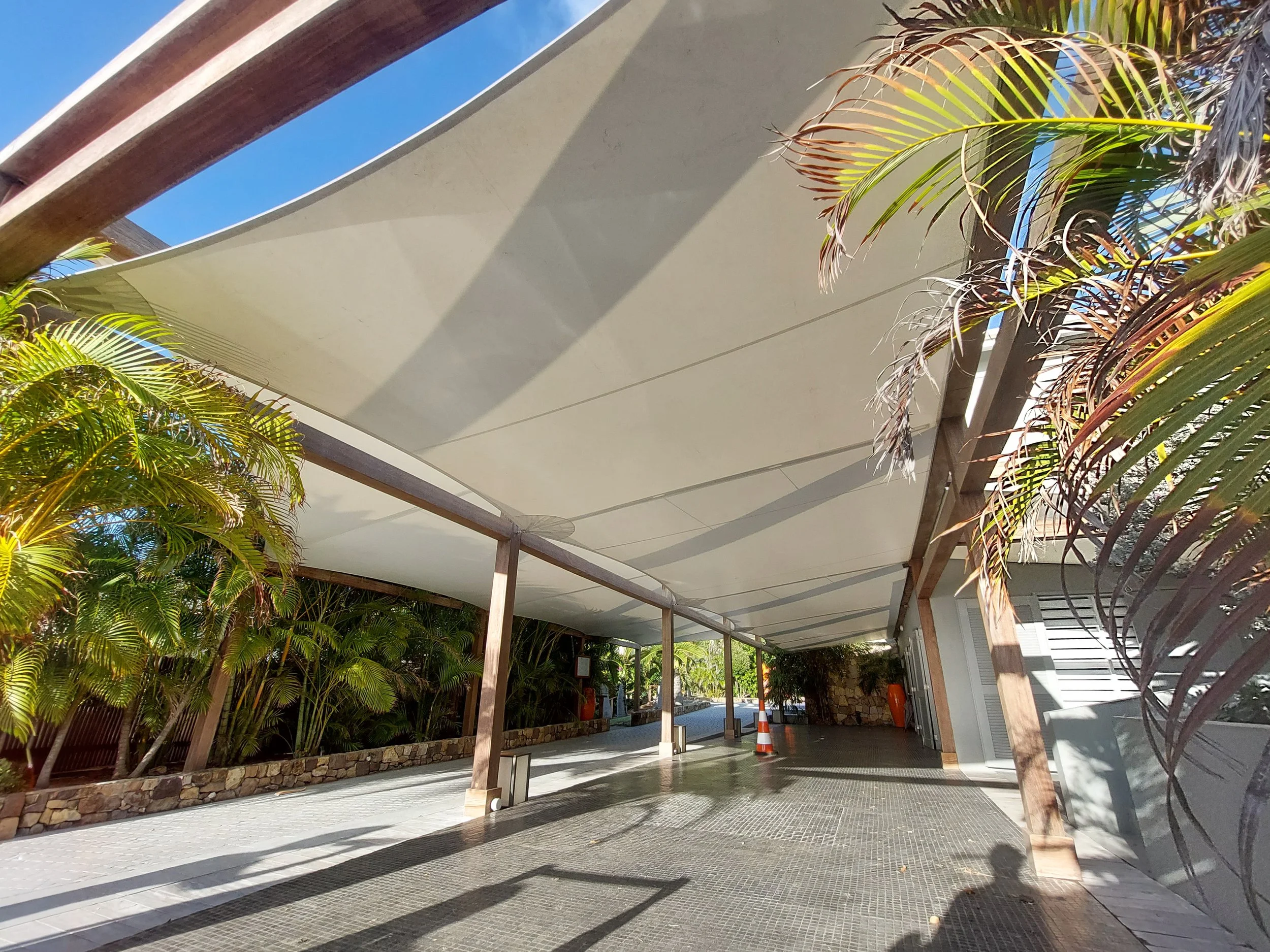 A modern outdoor area with a fabric canopy roof, tropical palm trees, stone planter bed, benches, and an orange traffic cone.