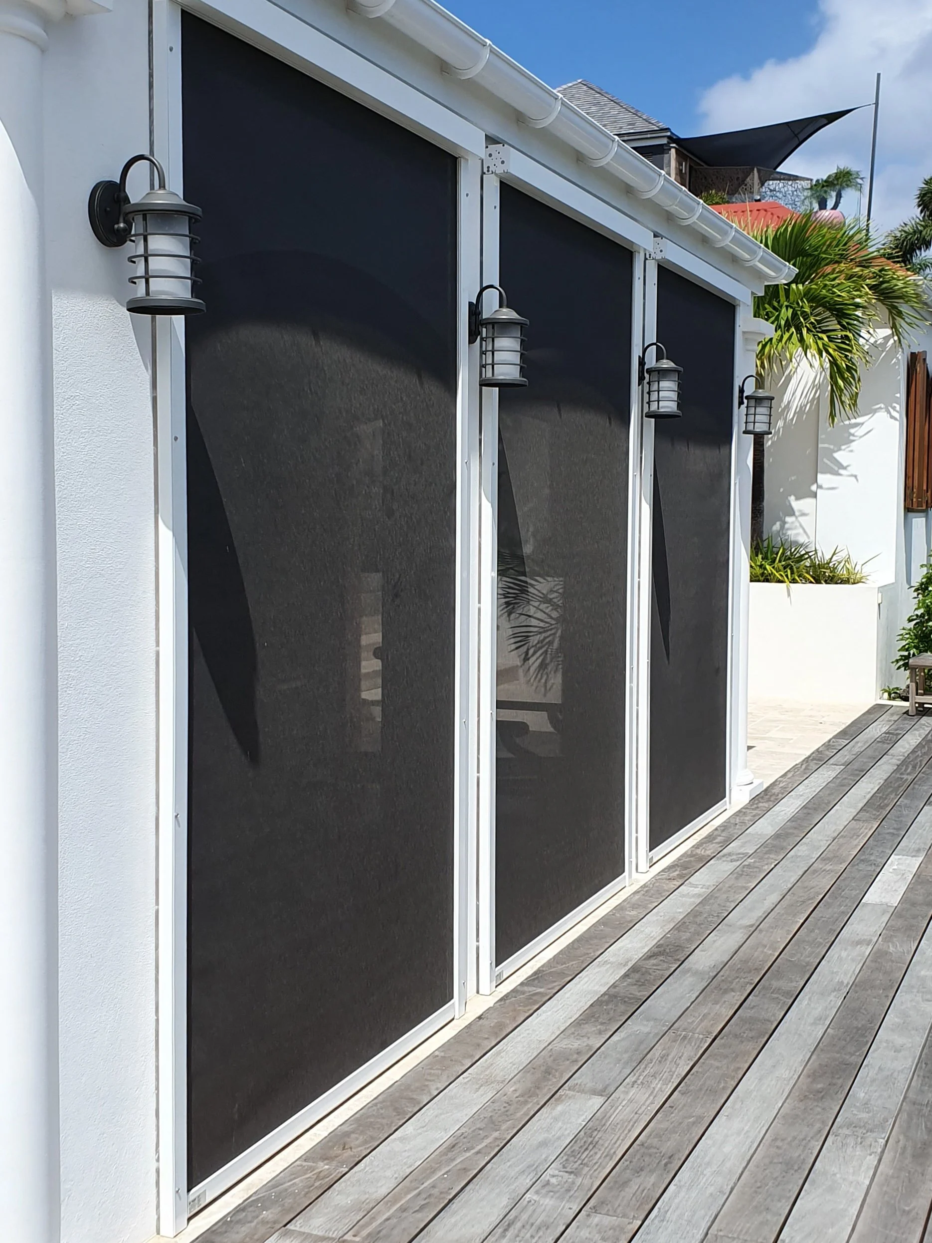 A patio with three black mesh sliding screens, three wall-mounted outdoor lanterns, a white wall, and wooden decking with a palm tree in the background.