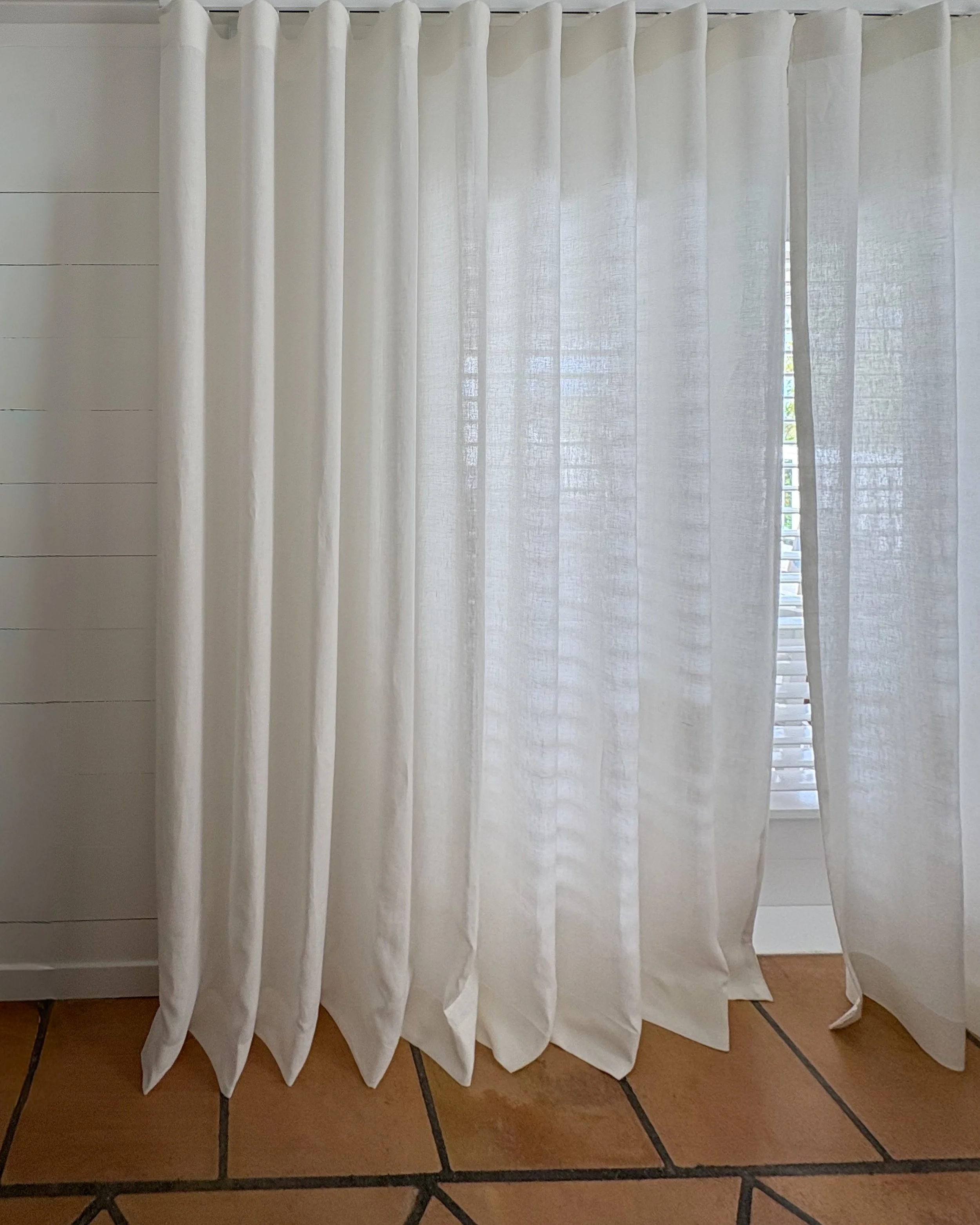 White curtains hanging in front of a window with blinds, partially opened, on a tiled floor.