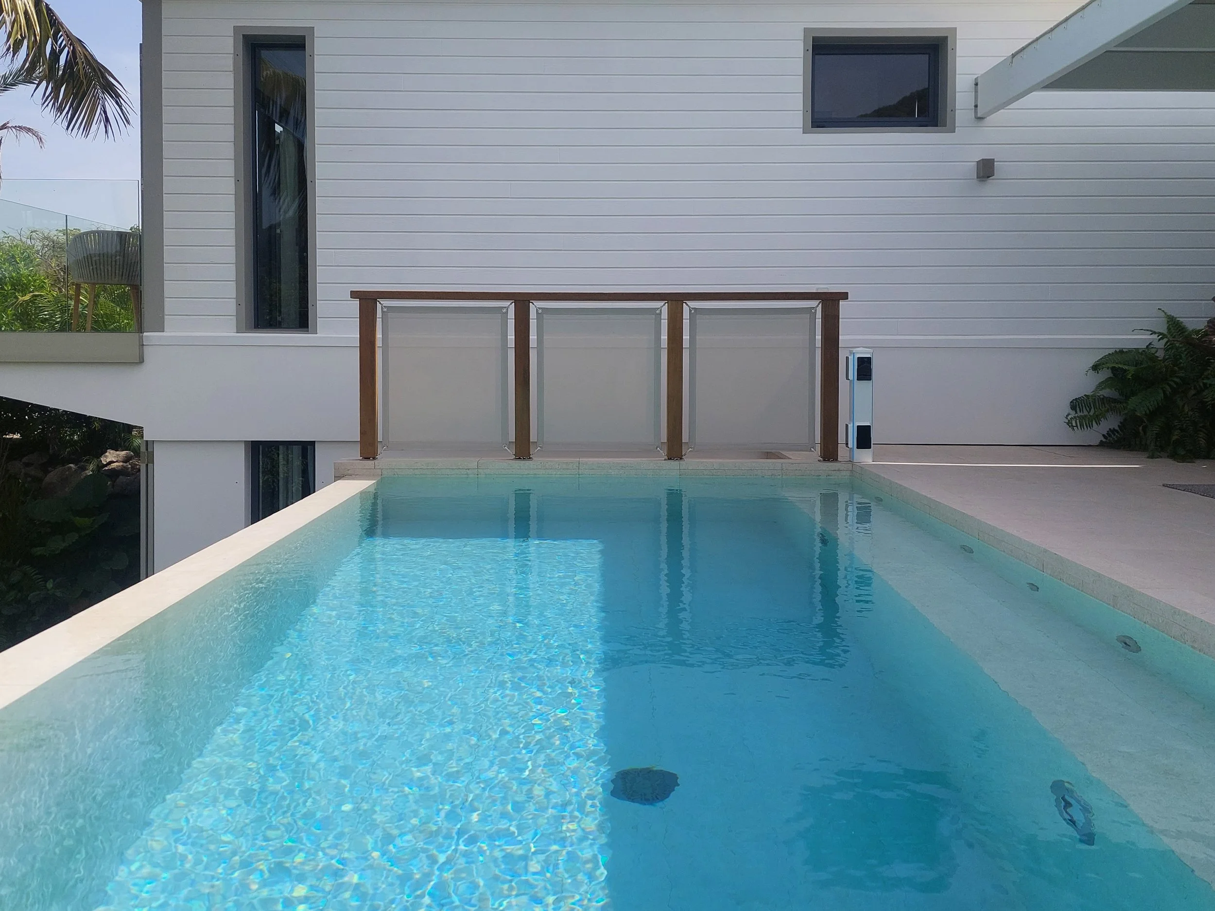 Outdoor swimming pool with clear blue water, surrounded by a white deck, with a modern white house in the background featuring windows, a balcony with a glass railing, and greenery.